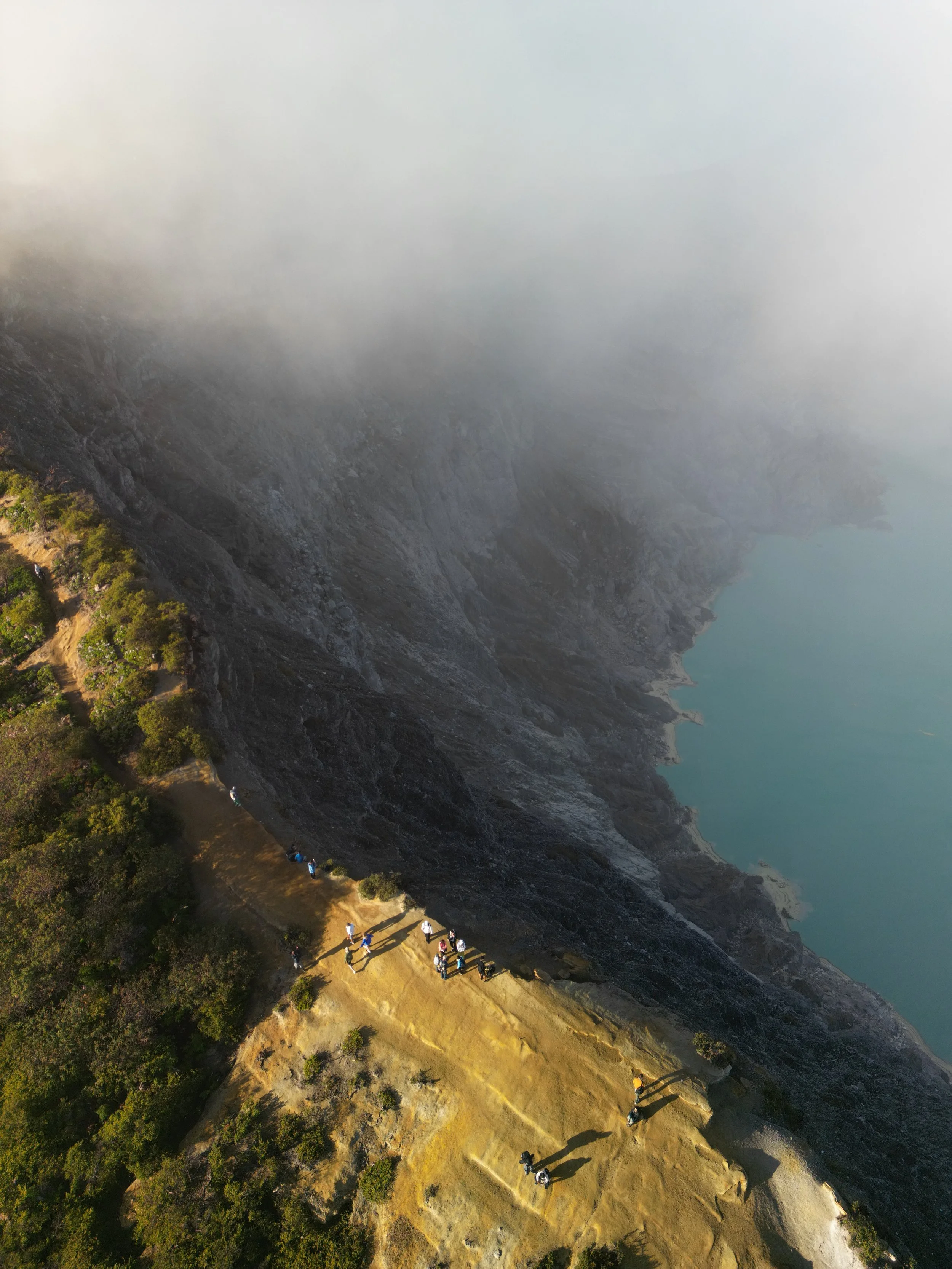 Aerial view of hikers walking along a cliffside trail above a volcanic lake at Ijen Volcano, Indonesia.
