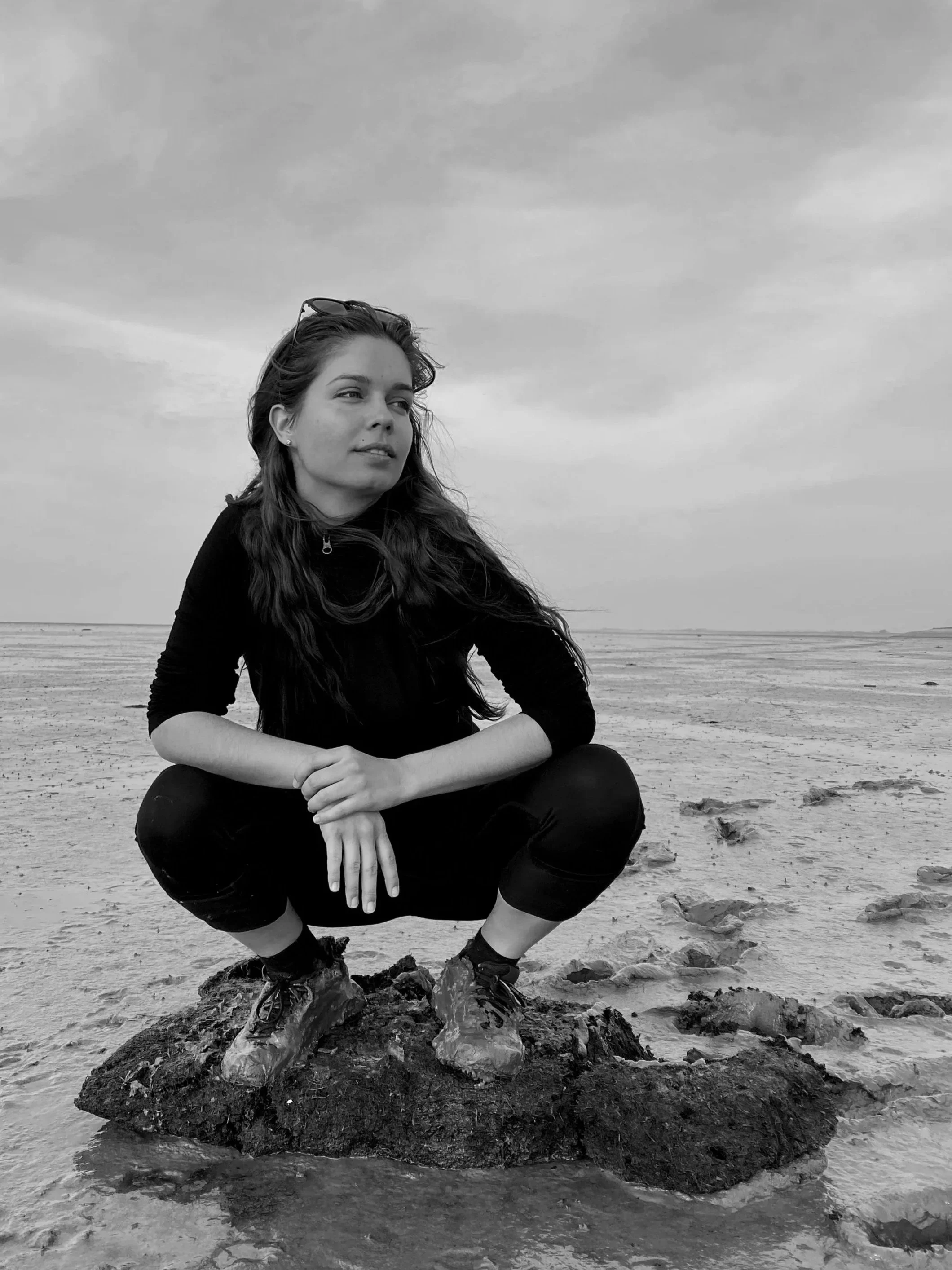 A woman with long hair, squatting on a rock on a beach, looking off to the side, wearing dark clothing and boots, with a cloudy sky in the background.