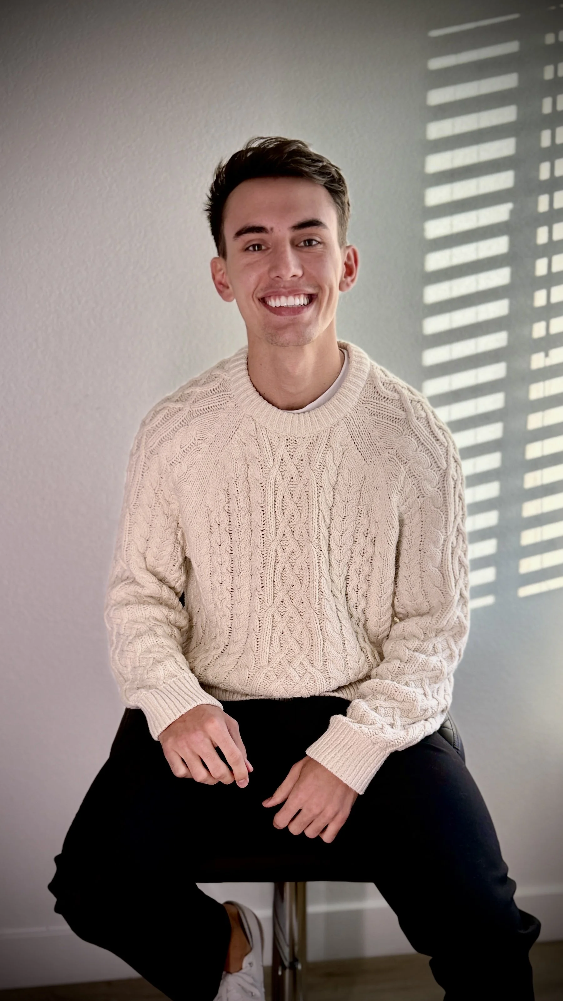 A young man with short dark hair and a bright smile, sitting on a black stool with his hands resting on his lap, wearing a cream-colored cable knit sweater and black pants, with light coming in from a window with horizontal blinds casting shadows on the wall behind him.