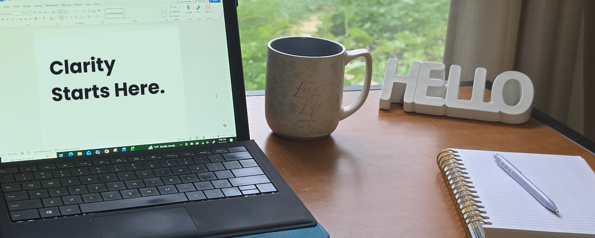 Picture of a workspace with a laptop coffee cup, notepad, pen, and 3D ceramic Hello sign. The laptop is open to a word processor and says Clarity Starts Here.