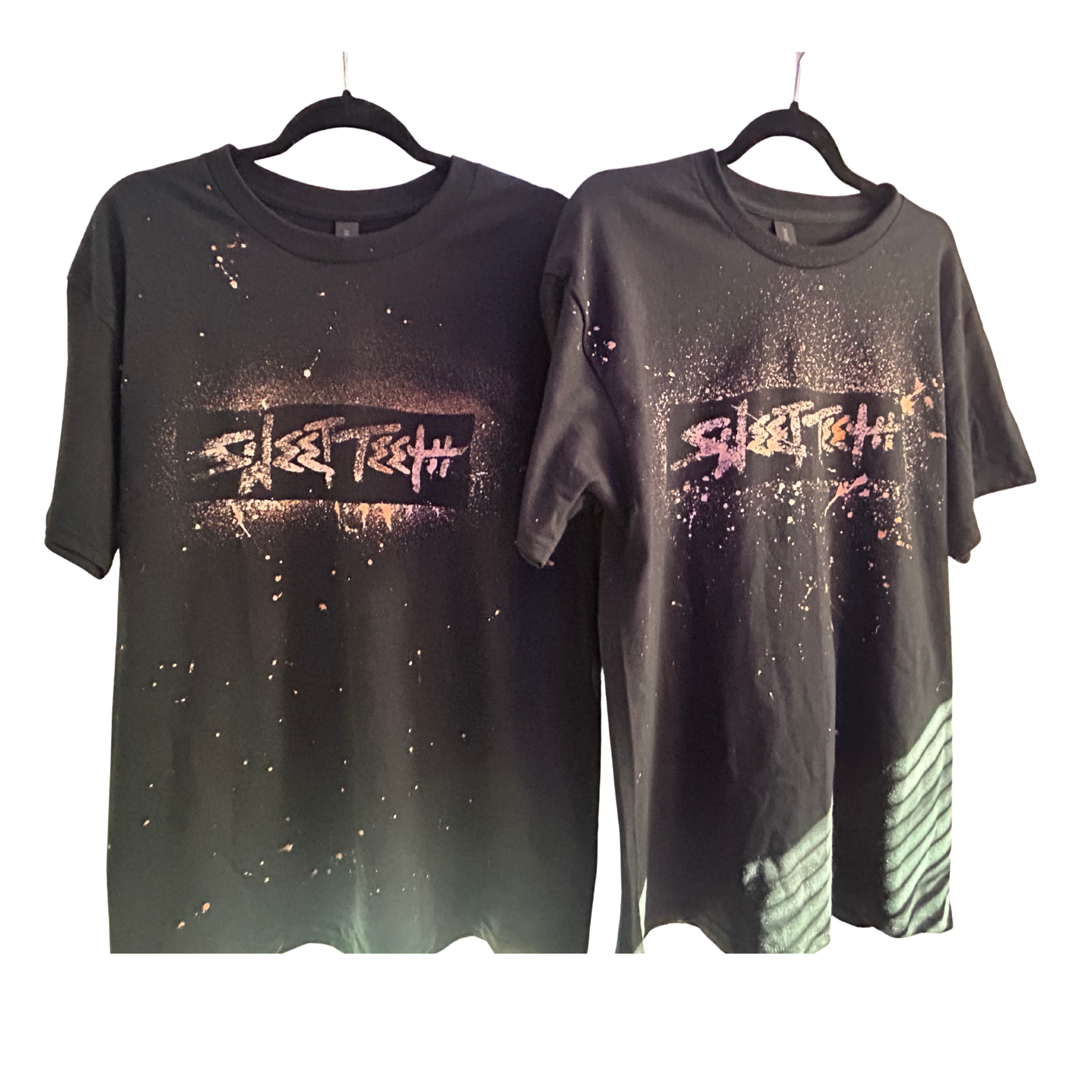 Two black T-shirts hanging on black hangers, featuring a splattered pink and white graphic design and stylized text, displayed against a black background.