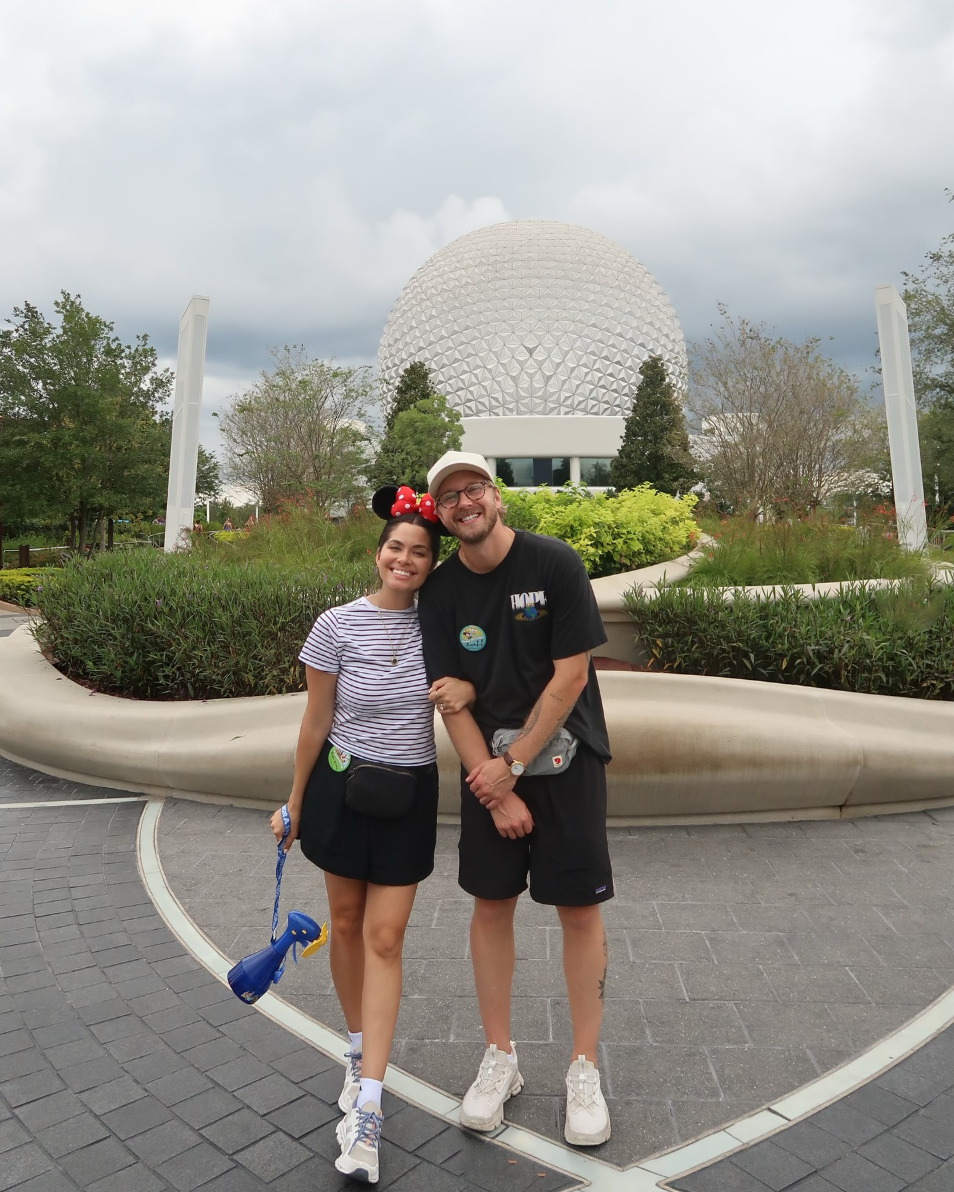 Two smiling people standing in front of the Epcot geodesic sphere at Disney World, with cloudy sky overhead and greenery around.
