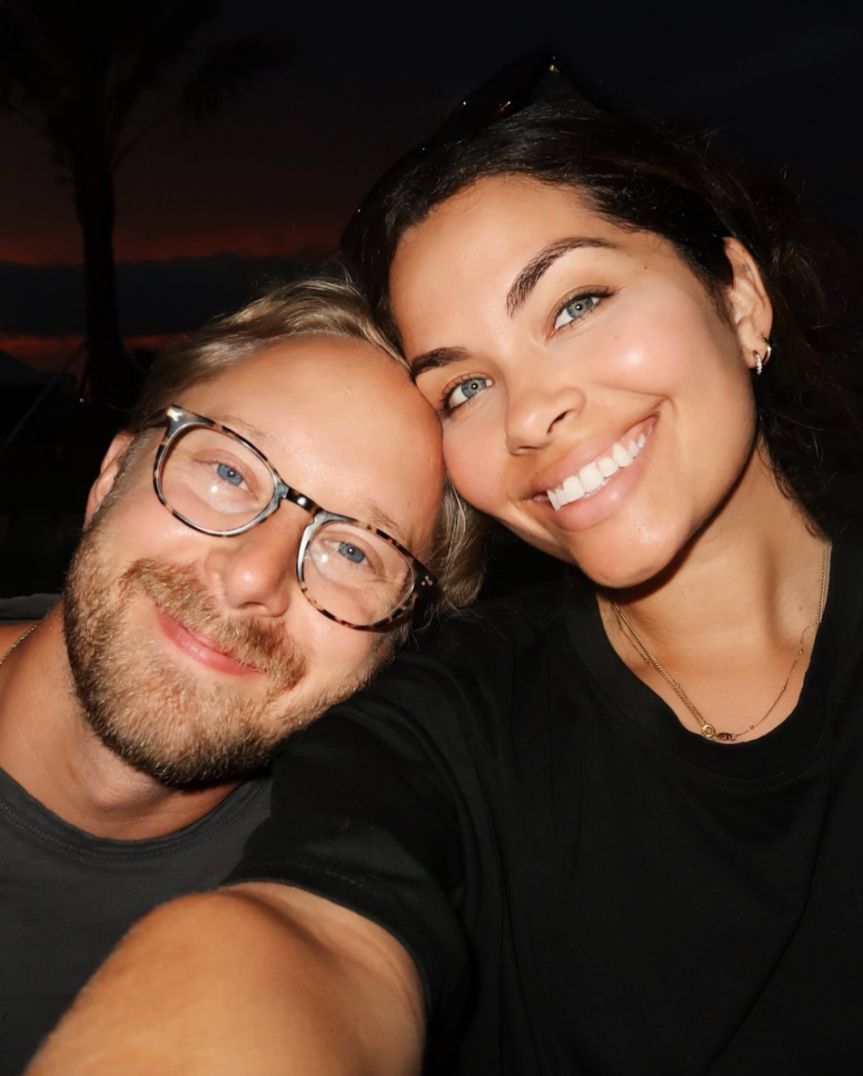 Close-up of a smiling man and woman taking a selfie together at sunset, with dark sky and a silhouette of a tree in the background.