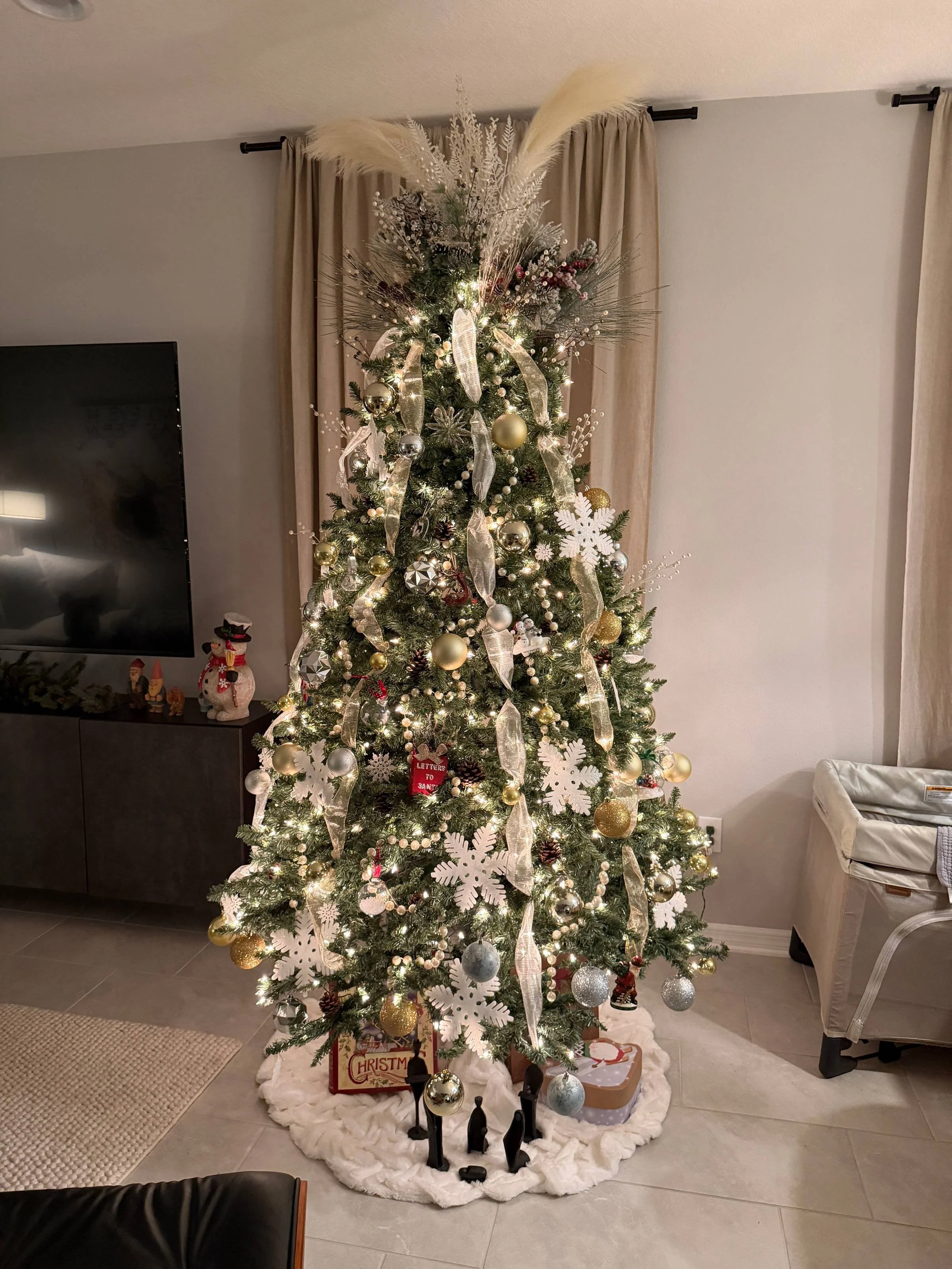 Decorated Christmas tree with white and gold ornaments, ribbons, and snowflakes, topped with pampas grass and lights, set in a living room corner.