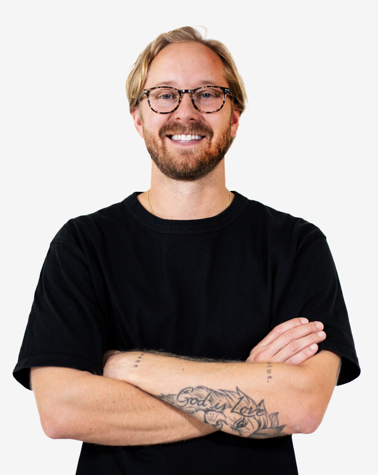 A smiling man with glasses, a beard, and tattoos on his arms, wearing a black T-shirt, standing with arms crossed against a white background.