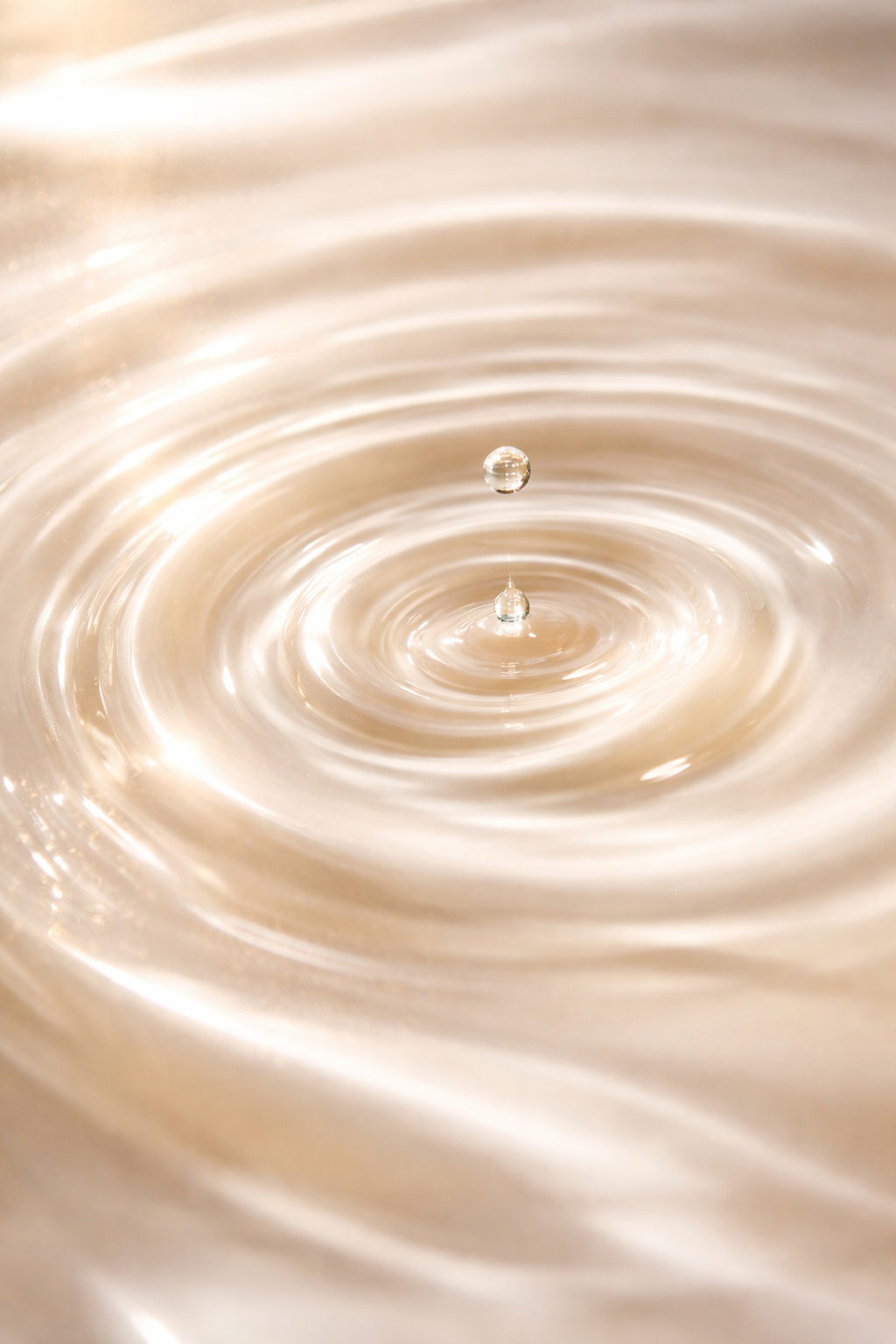 Close-up of a water droplet creating ripples on the surface of a liquid, with a bright, neutral background.