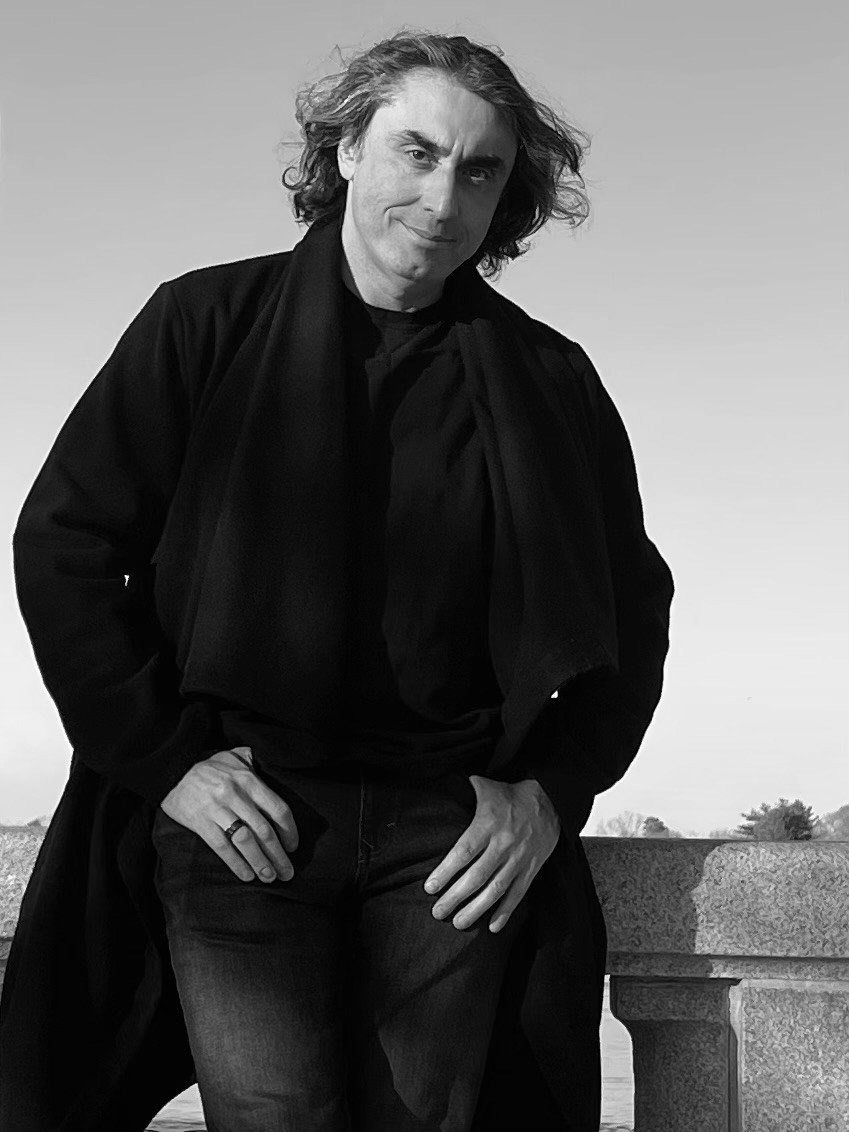 A man with long, wavy hair in black and white, wearing dark clothing, standing outdoors with hands in his pockets, slightly leaning against a stone railing.