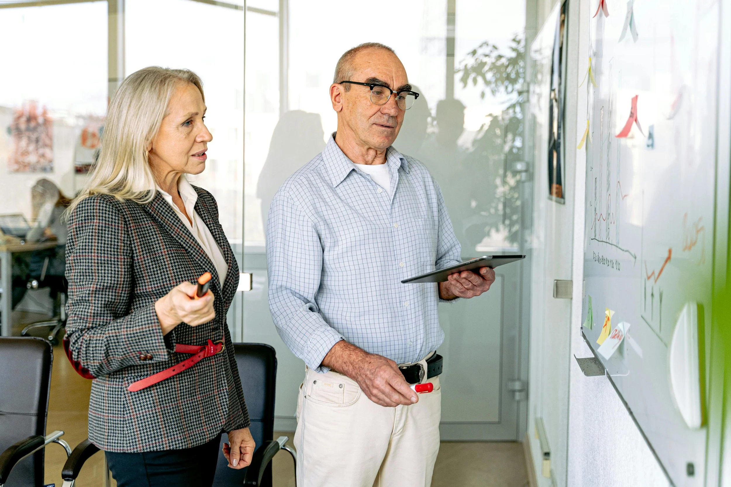 Two professionals, a woman and a man, stand in an office in front of a whiteboard with charts and notes, engaged in a discussion.