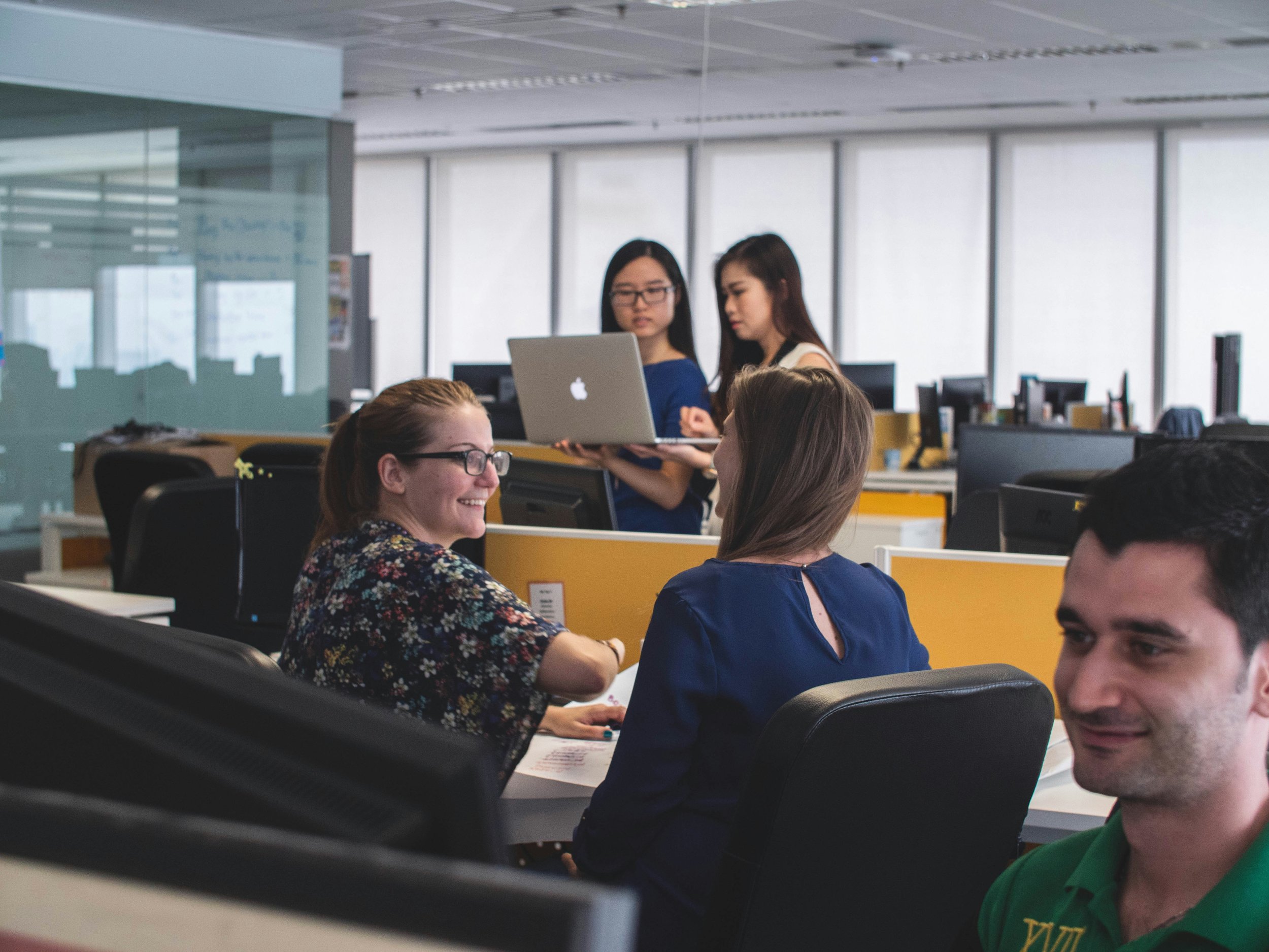 Office workers in a modern open-plan office with desks and computer monitors, engaging in a discussion and working on computers.