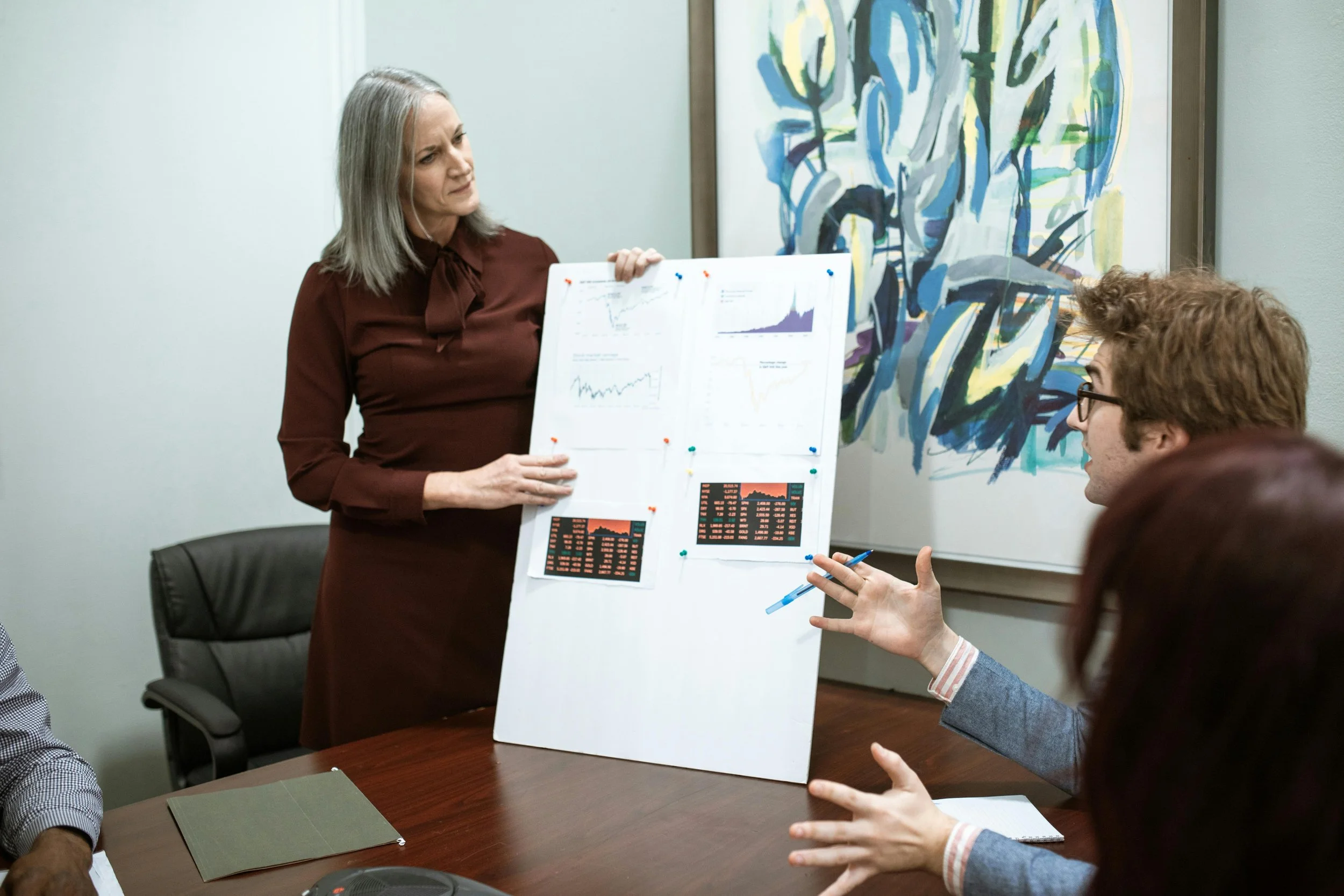 A woman in a burgundy dress presenting a white poster board with charts and graphs to a group in a meeting room. Two men and one woman are seated at the table, engaging in discussion.