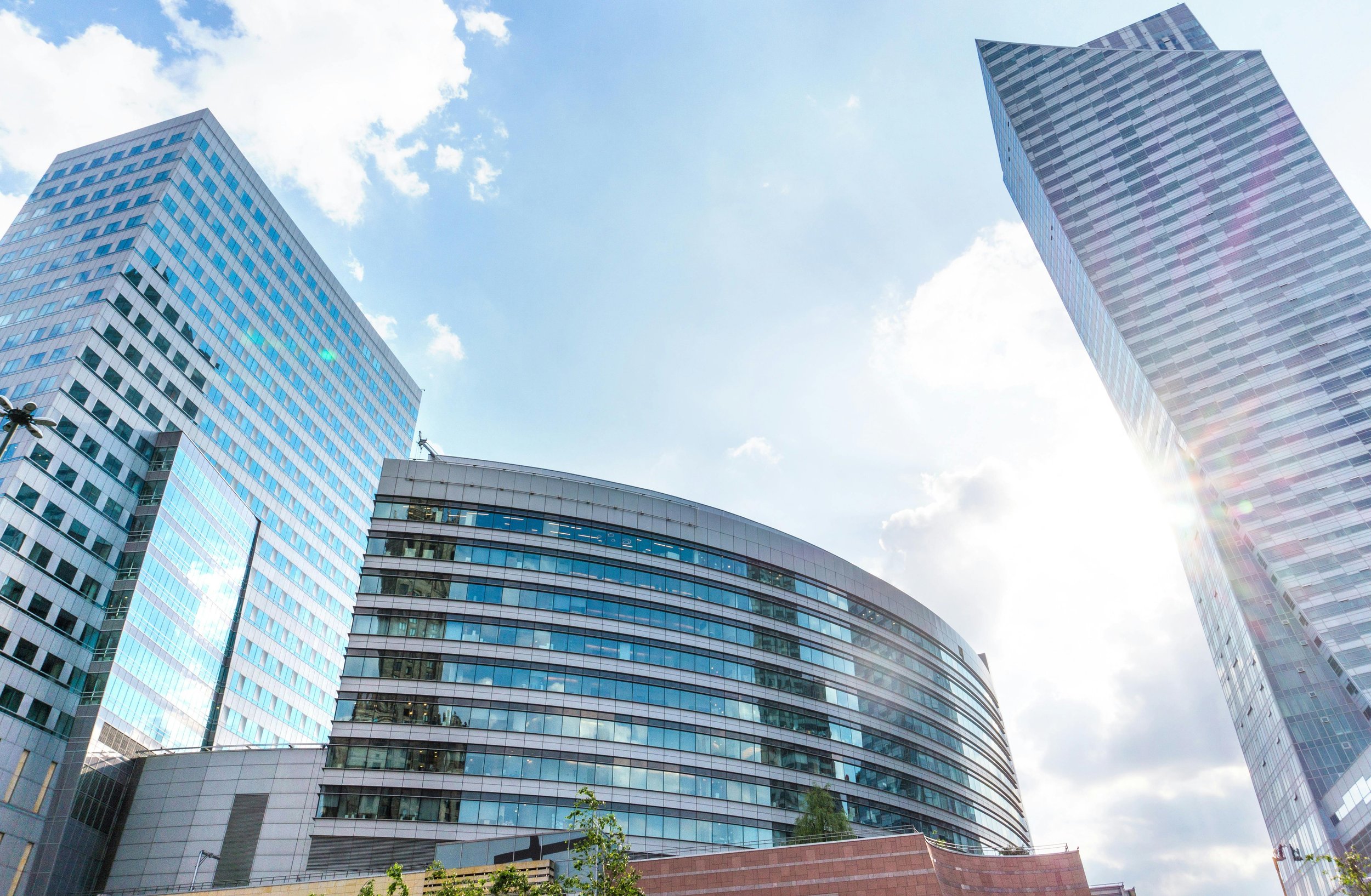 A cityscape with three tall, modern glass buildings under a partly cloudy sky with sunlight.