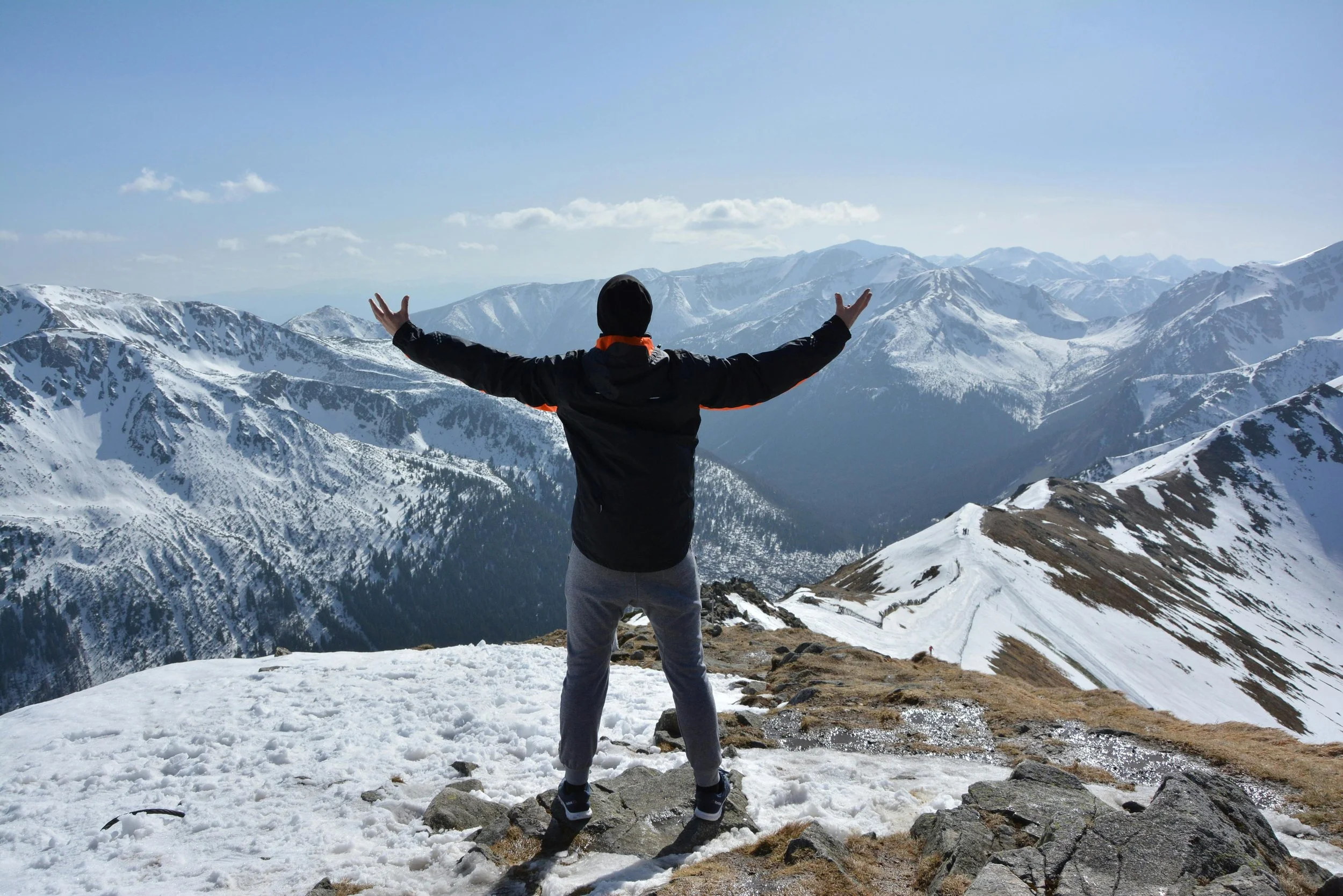 Person standing on snowy mountain ridge with arms outstretched, overlooking snow-capped mountain range under clear blue sky.