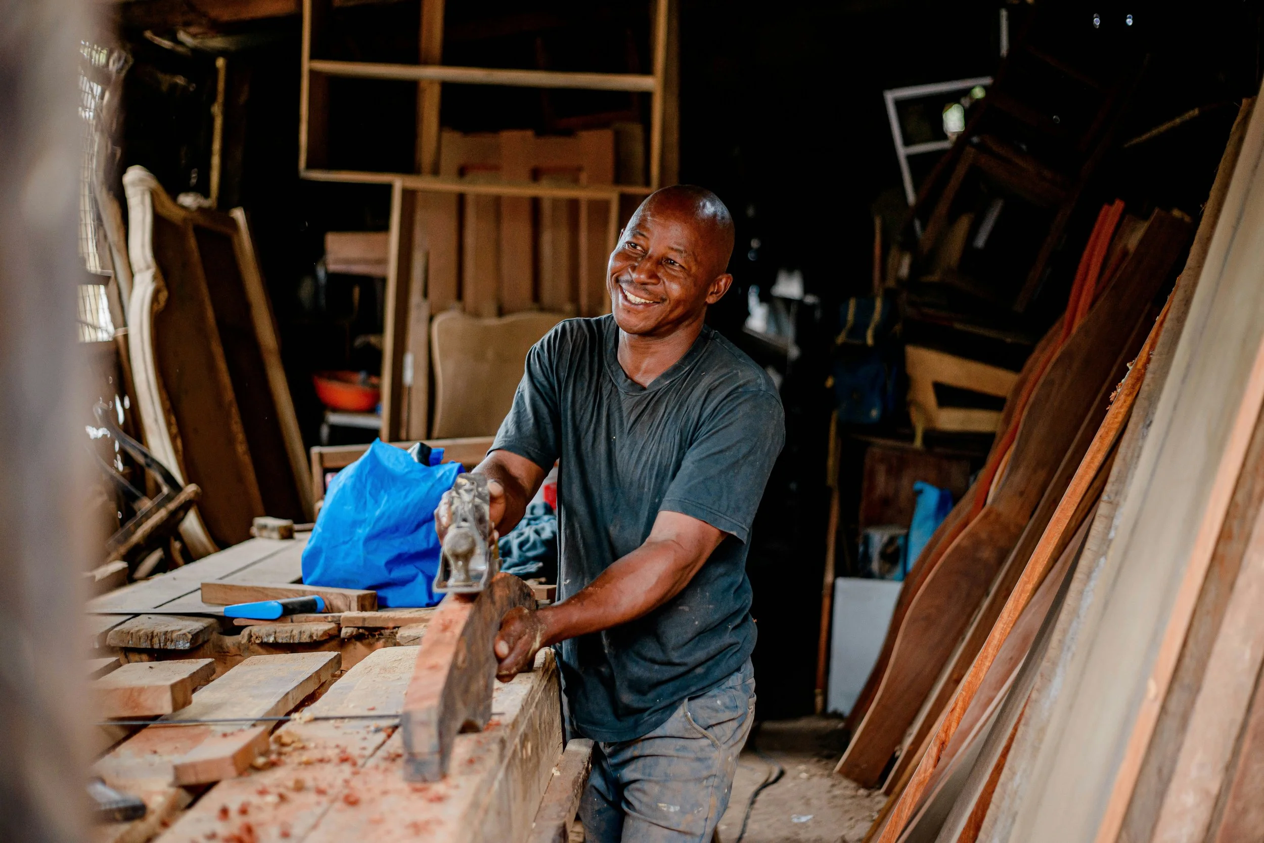 A smiling man working with a hammer in a woodworking shop, surrounded by wooden furniture and supplies.