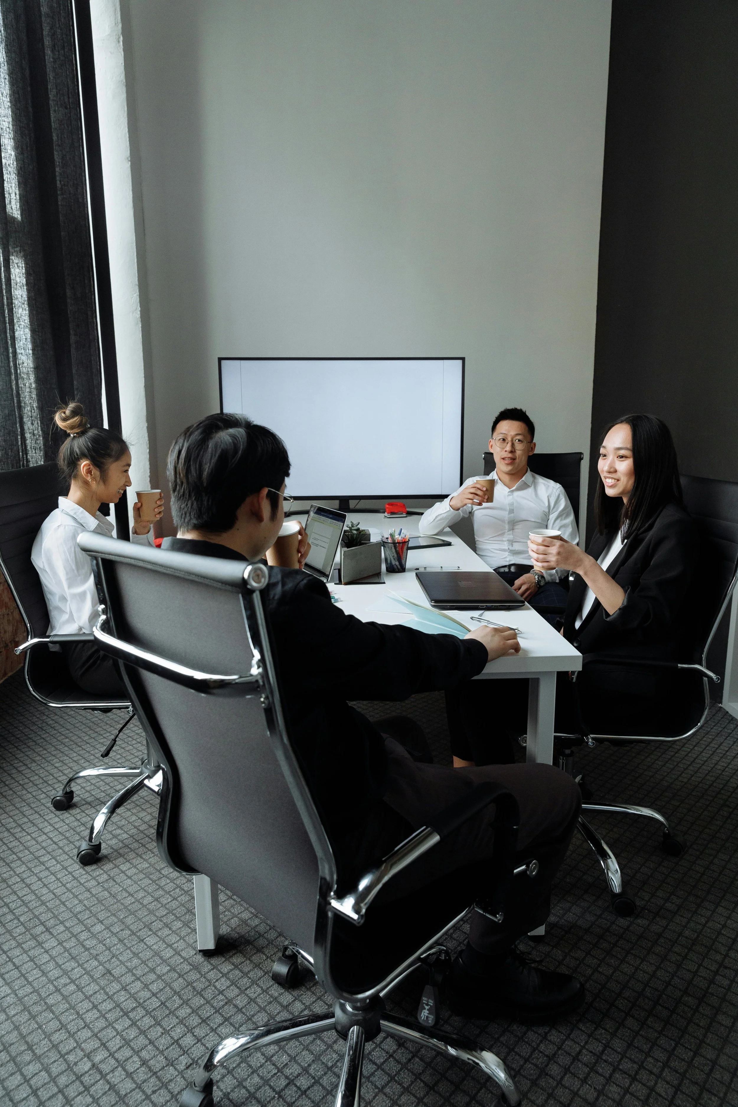 Four professionals in business attire having a meeting in a conference room, sitting around a table with laptops and documents, with a large monitor on the wall and coffee cups in hand.