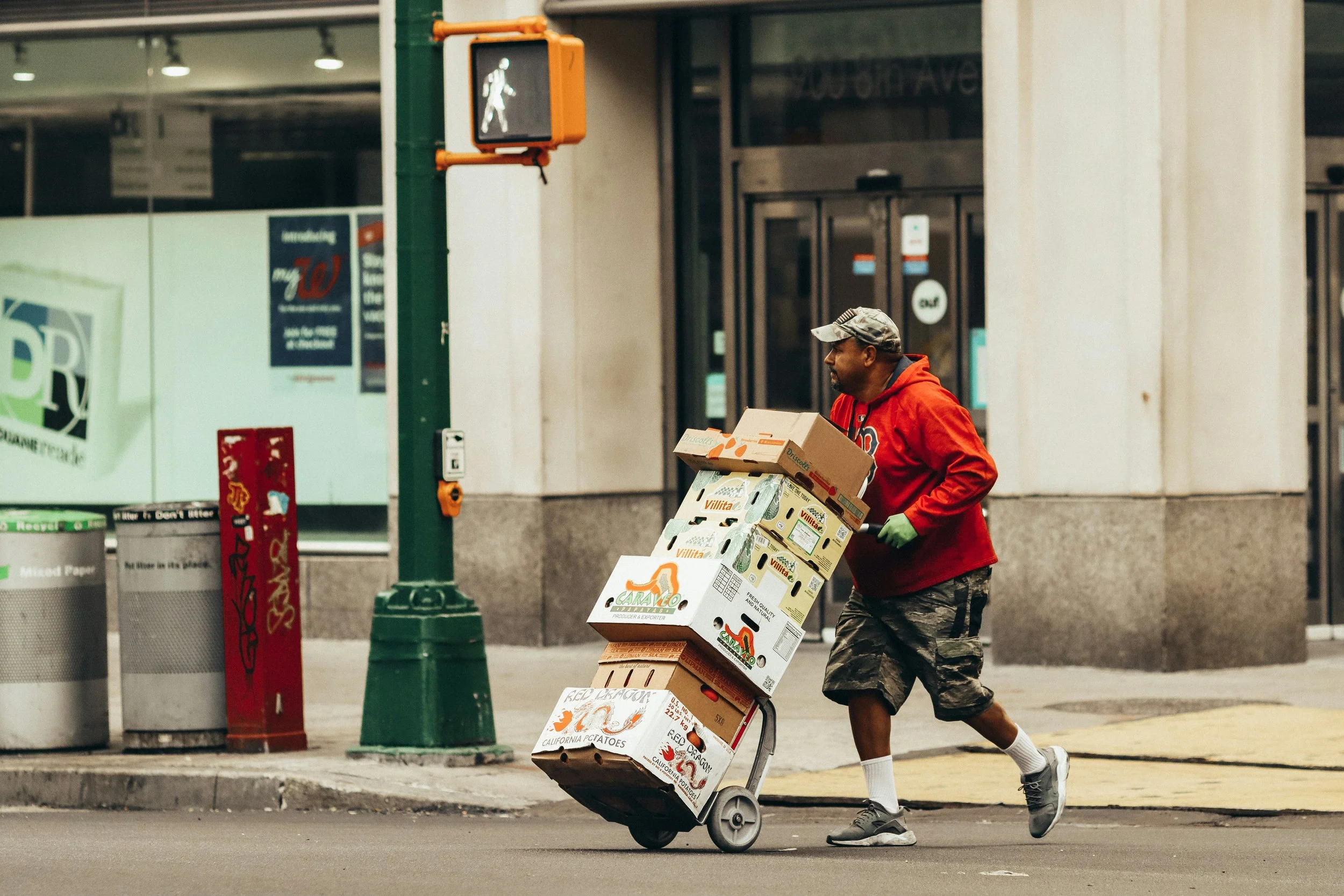Man pushing a hand truck stacked with cardboard boxes across a city street
