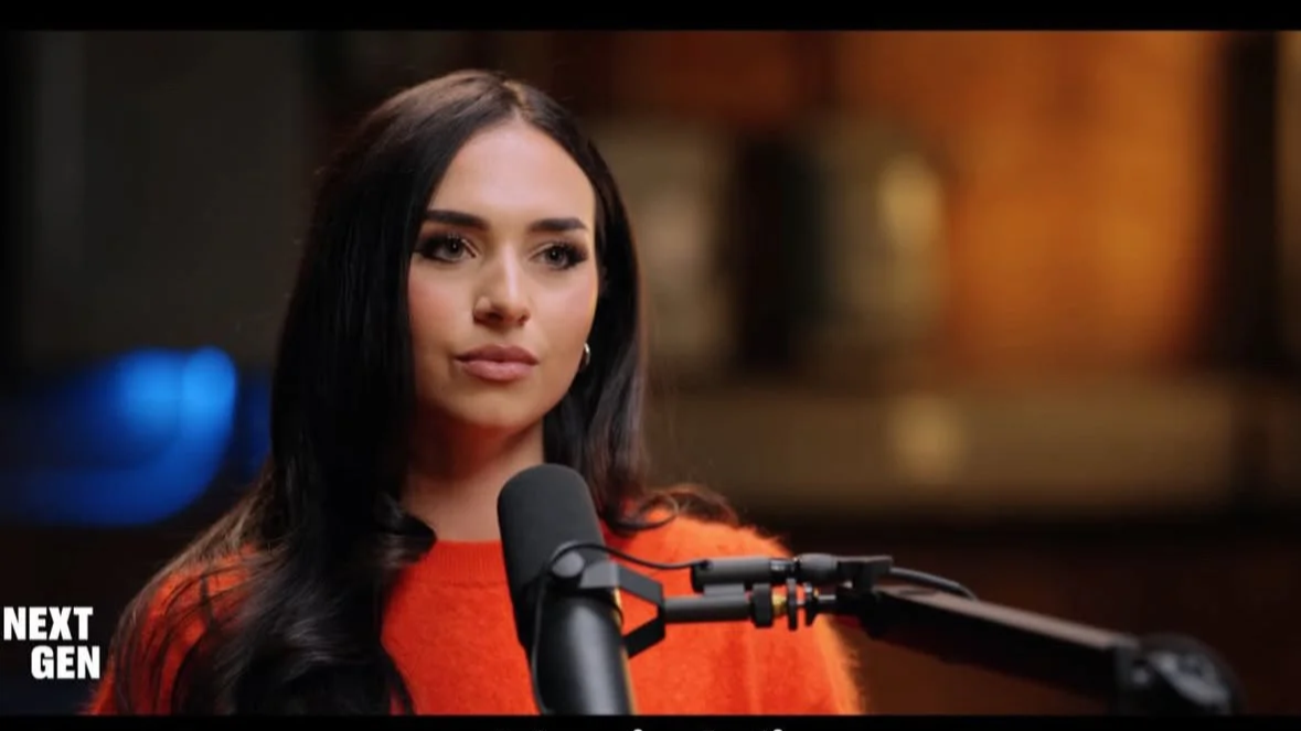 Gia Chacon wearing an orange sweater speaking into a microphone on a stand, with a blurred background.