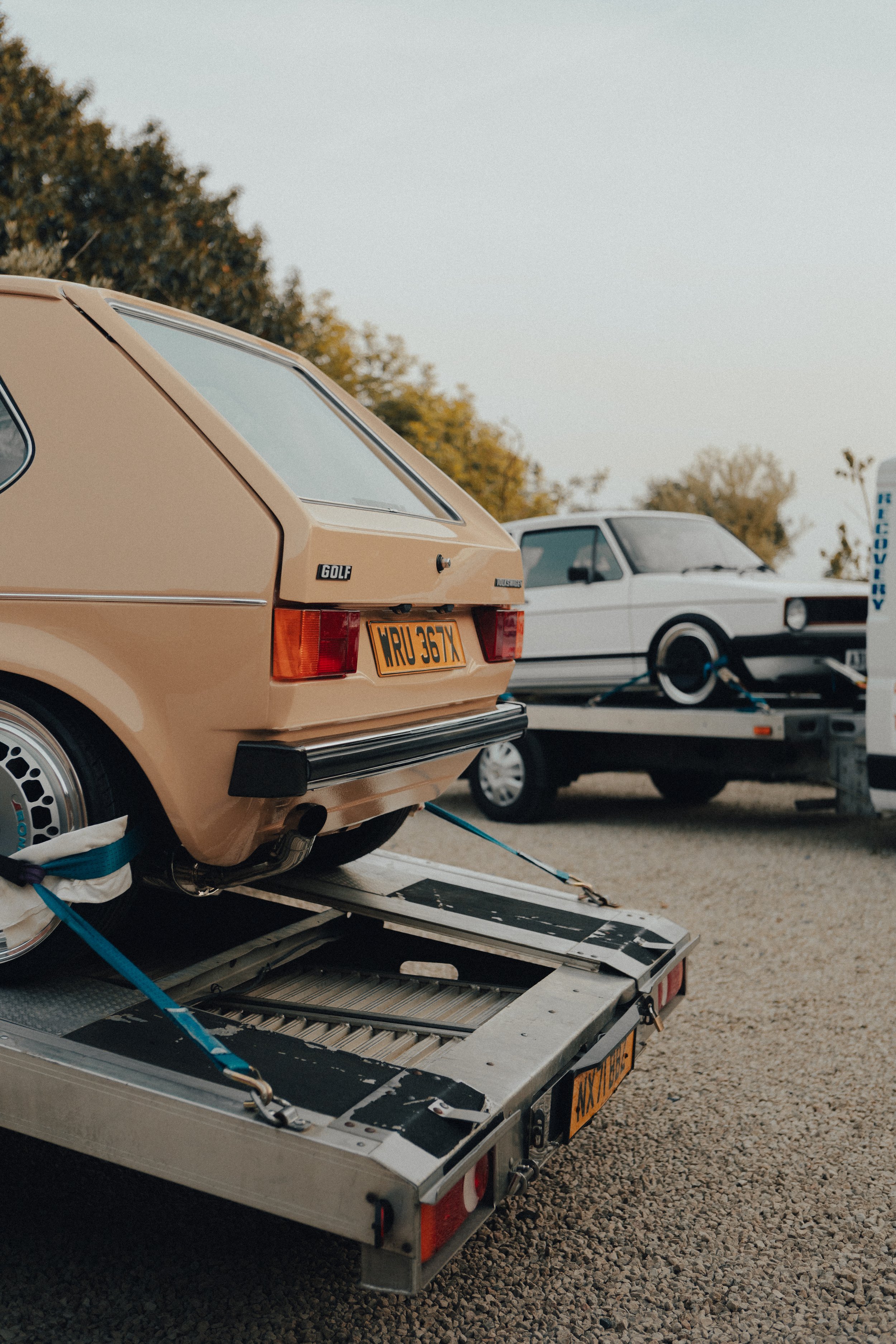 A vintage beige hatchback car on a trailer, secured with straps, with a white car on a flatbed trailer in the background at an outdoor parking lot during daytime.