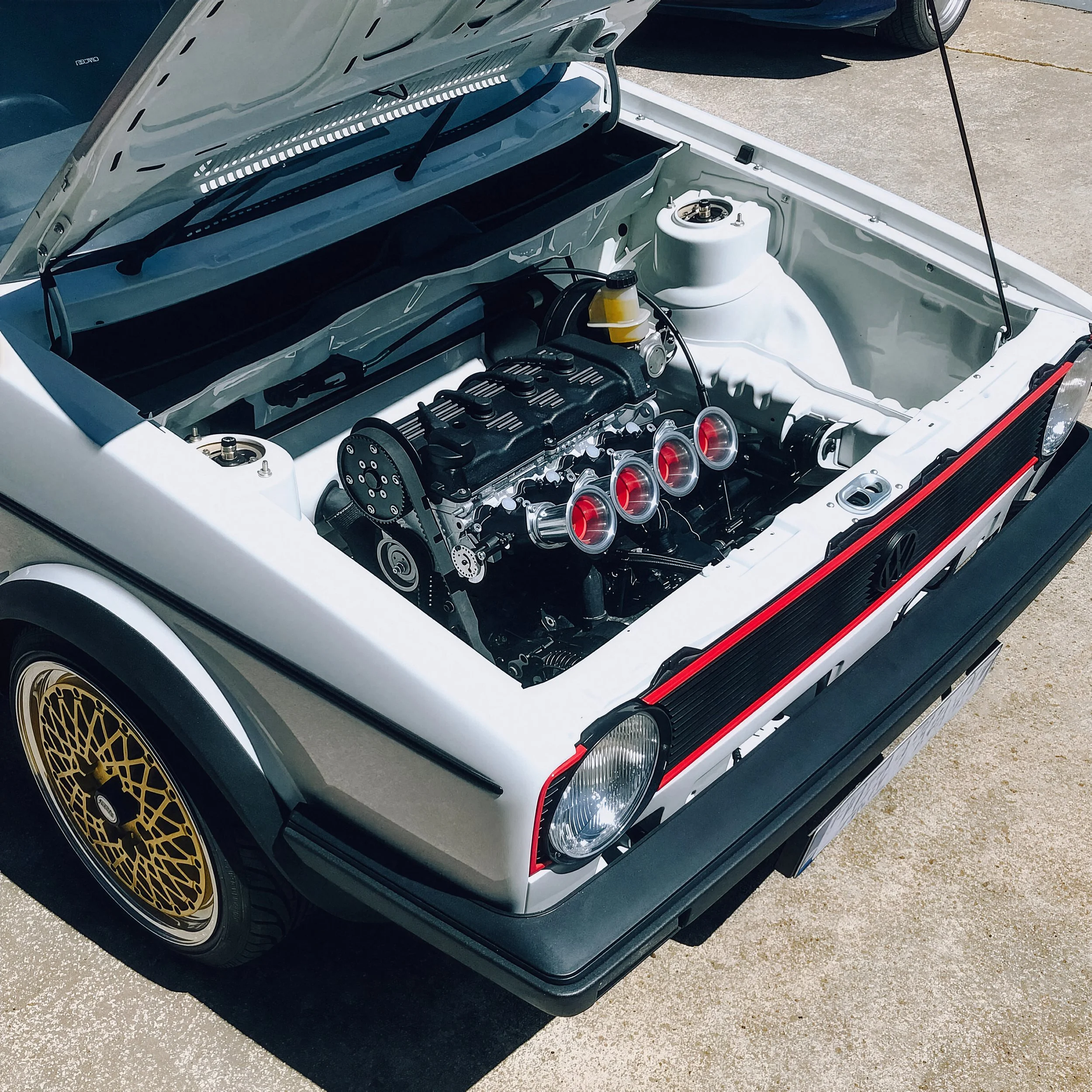 A white vintage car with an open hood revealing a high-performance engine with four red-tinted intake tubes. The car has gold BBS wheels and is parked on a concrete surface.