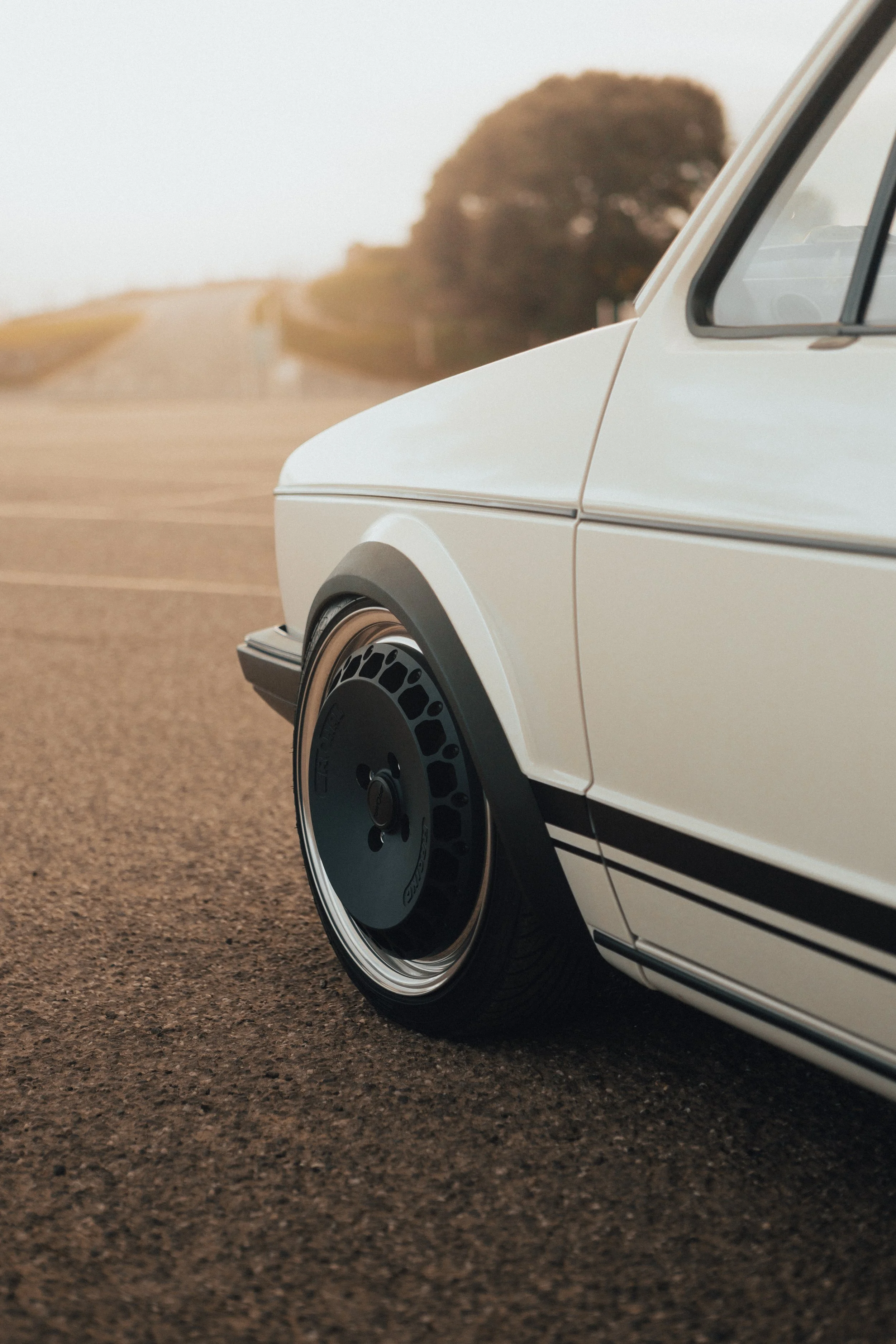 Close-up of the front left side of a vintage white car with black accents, parked on a dirt road, with trees and a distant hill in the background.