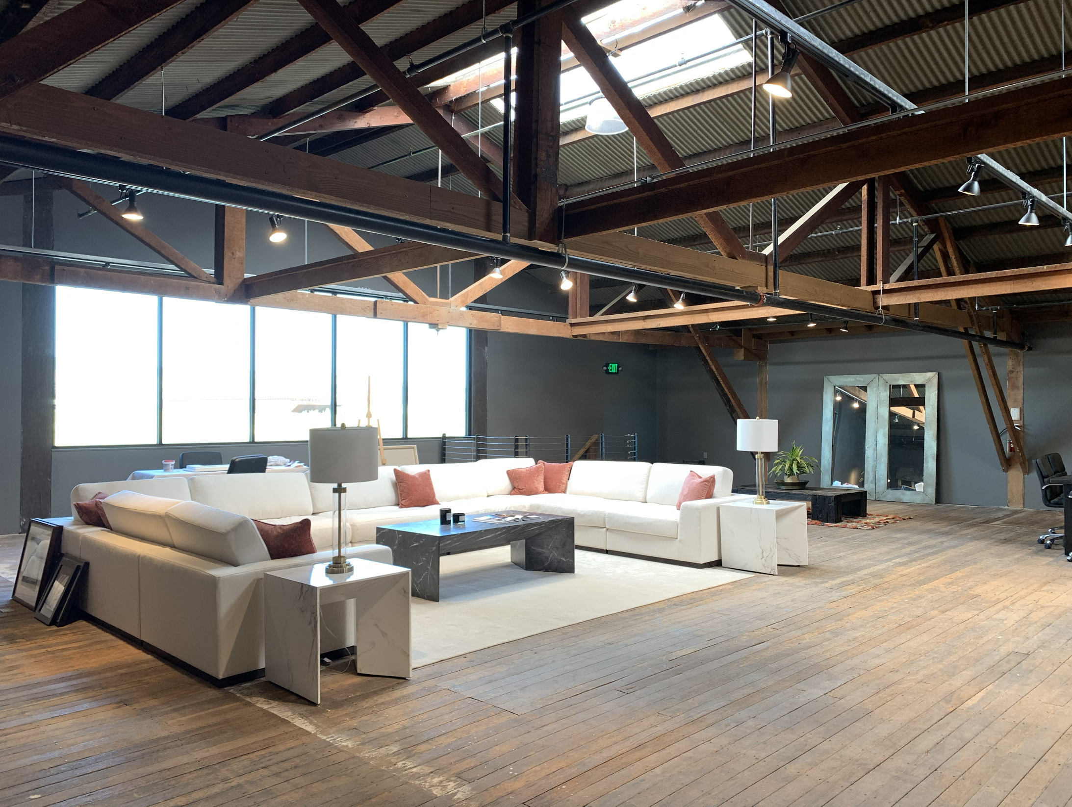 Spacious loft-style room with a large white sectional sofa, black marble coffee table, side tables with lamps, and decorative pillows. Large windows let in natural light, and wooden beams and floors add rustic charm. There is a mirror leaning against the wall and a potted plant near a black bench.