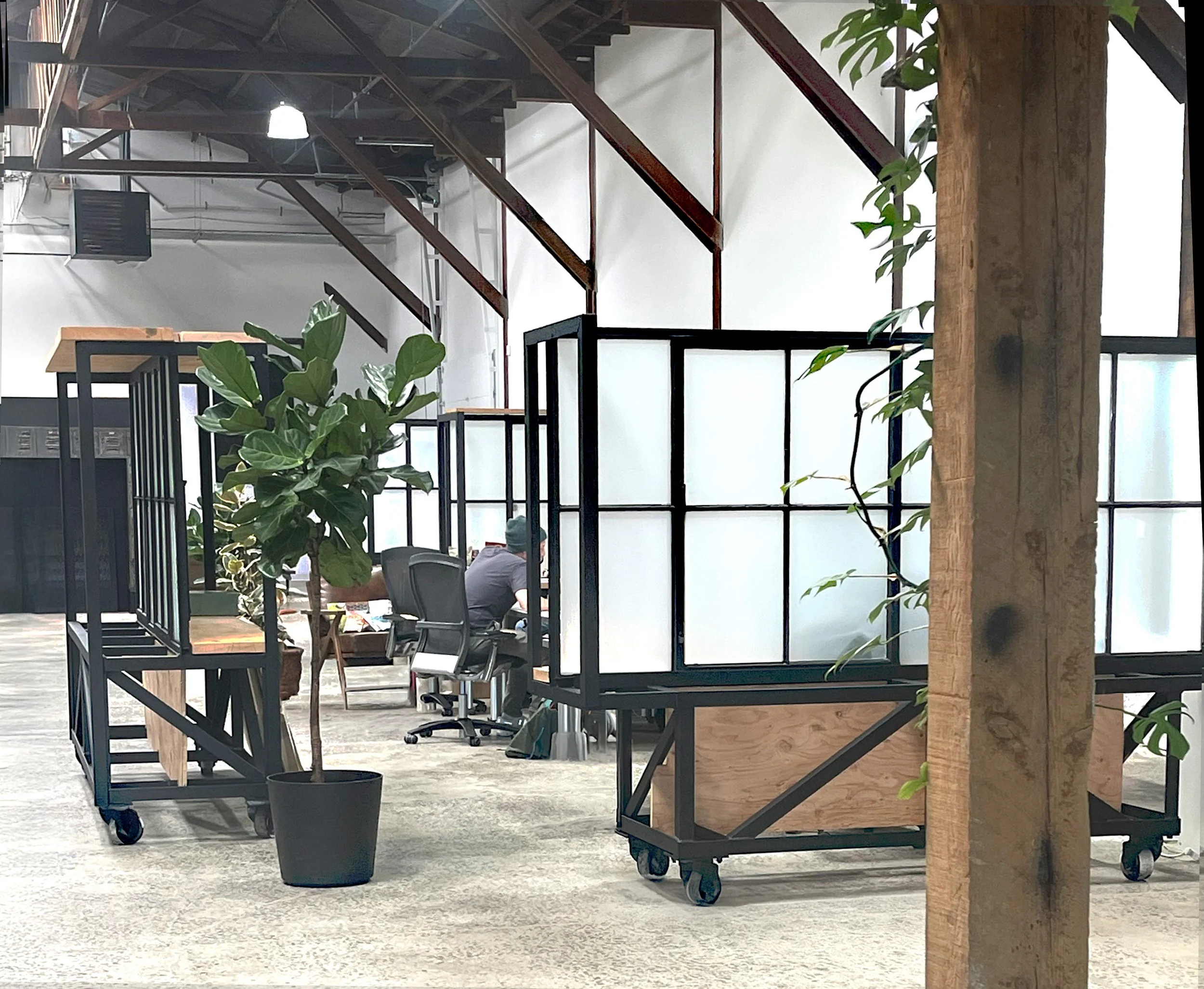 An industrial-style office space with wooden and black metal furnishing, potted plants, and a person working at a desk in the background.