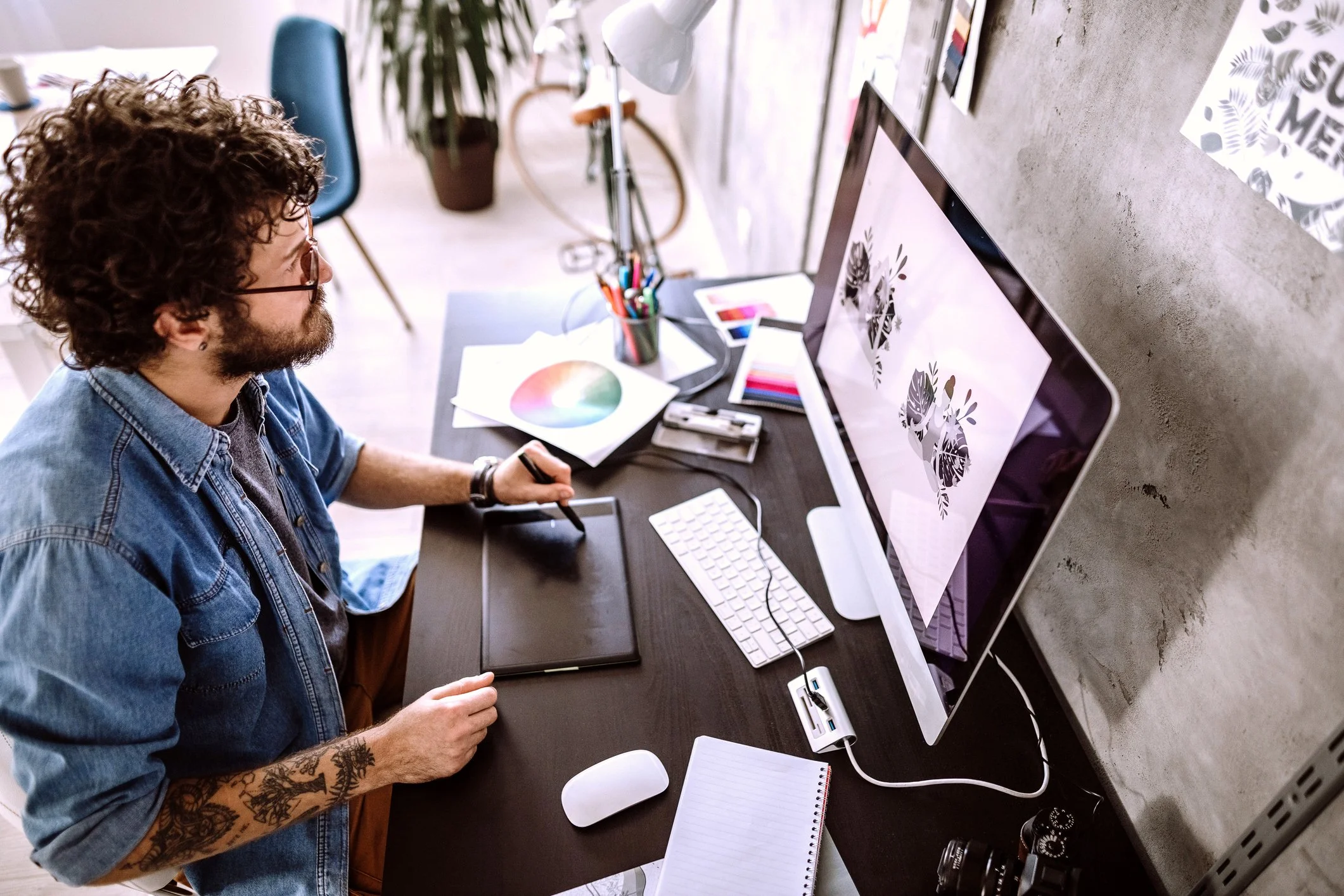 Man working at a digital art workstation with a large computer monitor, graphic tablet, color palette, color swatches, and creative supplies in a modern workspace.