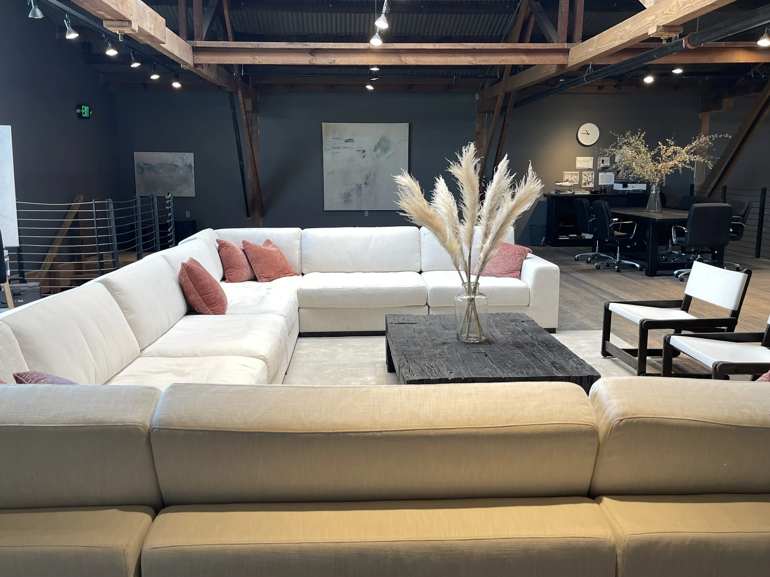 Modern living room with a large white sectional sofa, pink throw pillows, a rustic wooden coffee table with a glass vase of pampas grass, black and white chairs, and a workspace area with a table, chairs, and decorative plant in the background.