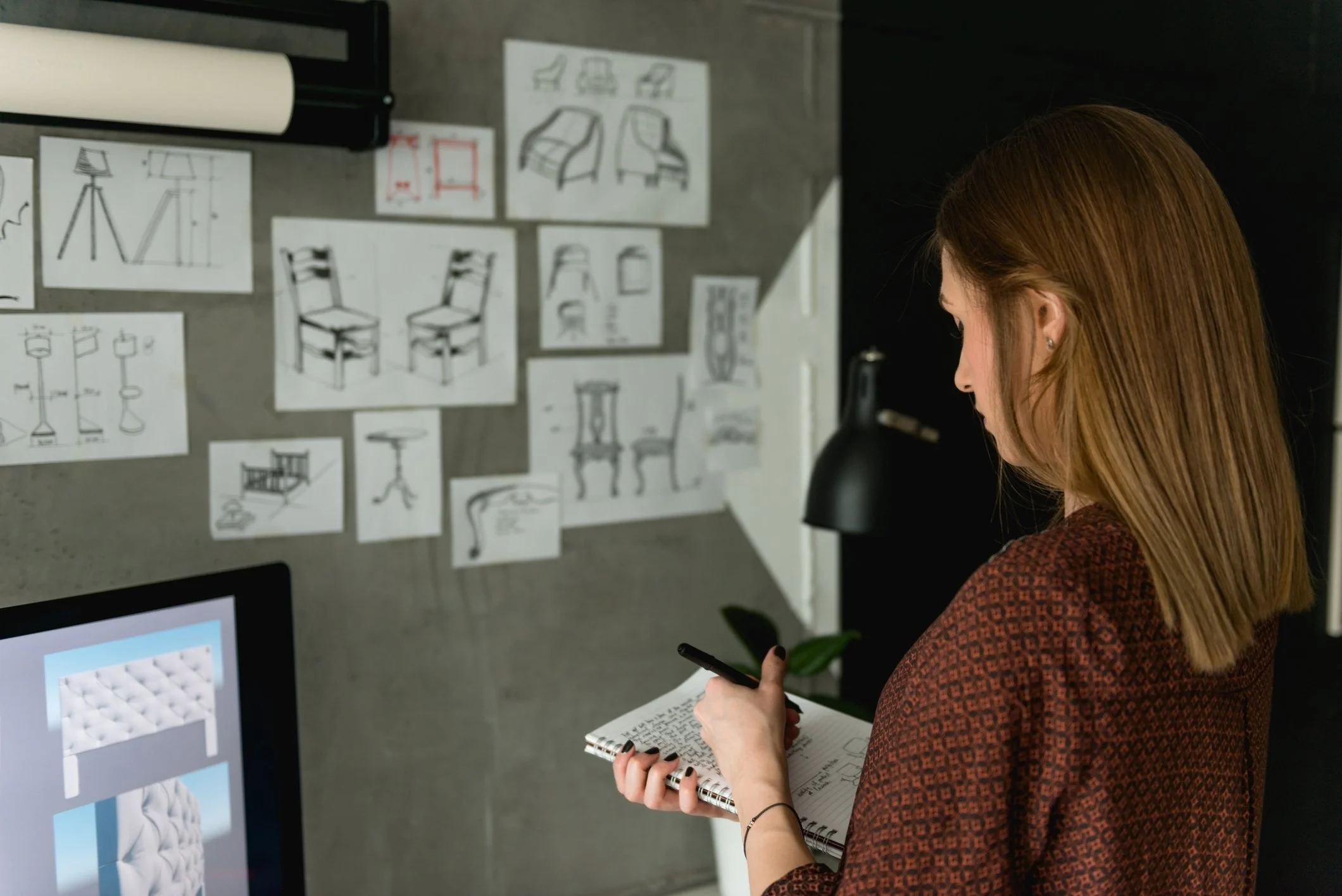 A woman with shoulder-length brown hair and a checkered blouse takes notes while standing in front of a wall covered with sketches of furniture designs.