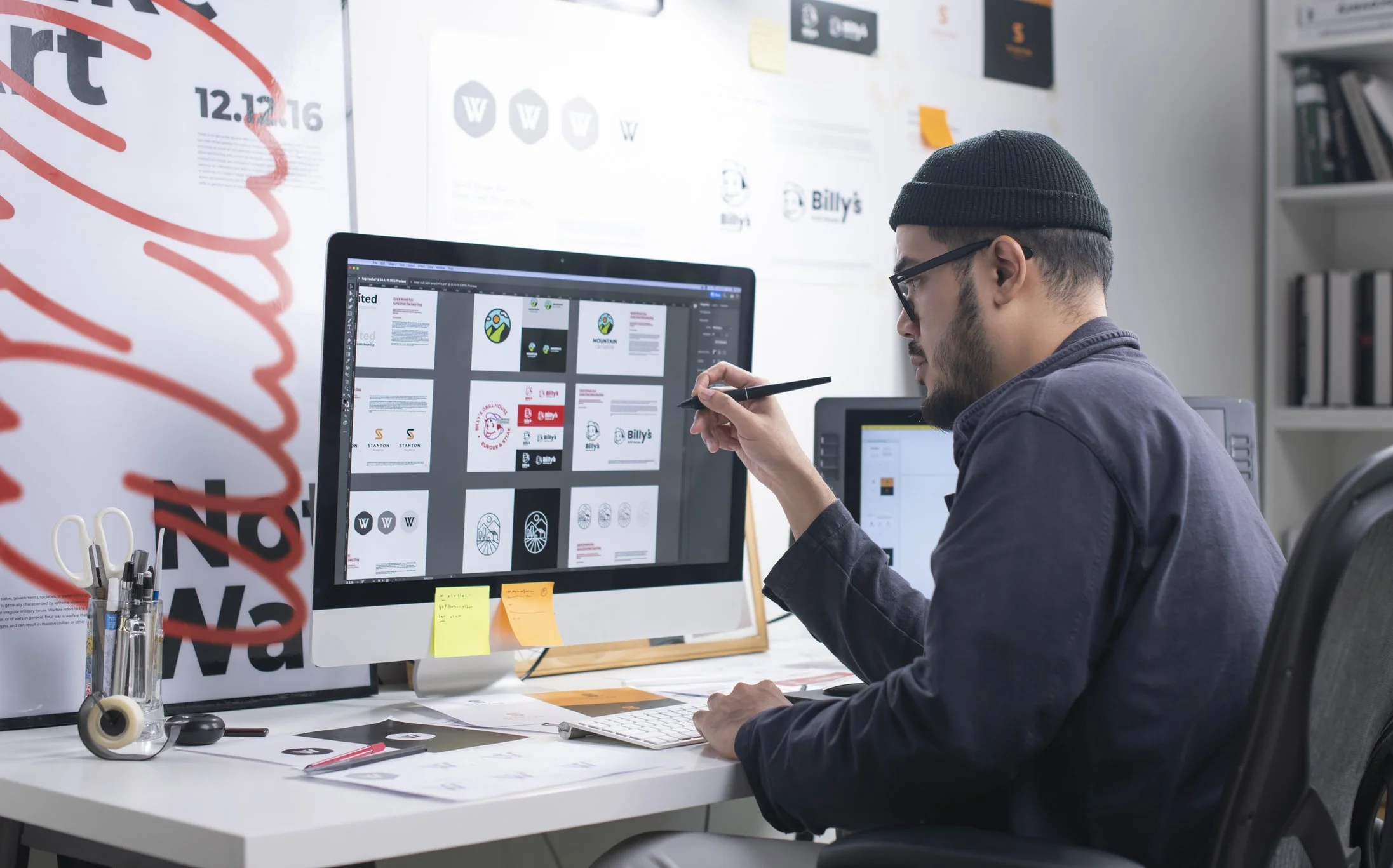 Man designing graphics on a computer in an office with design materials and sticky notes.