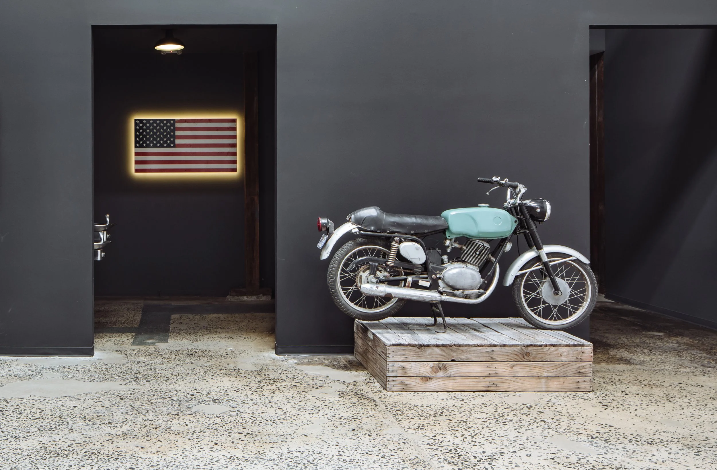 A vintage motorcycle displayed on a wooden platform next to a black wall with a backlit American flag, situated in a modern interior space.
