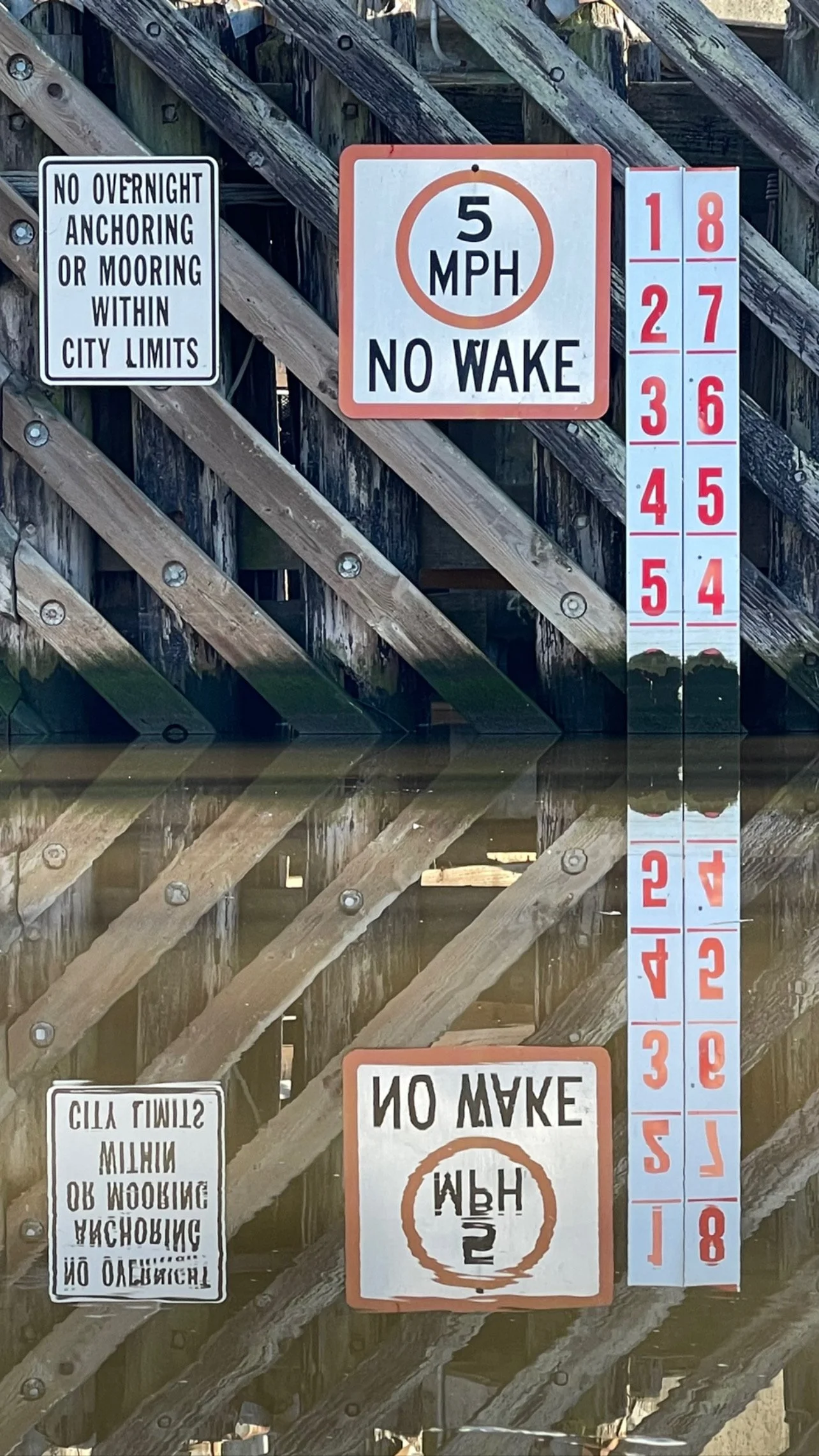 Signs on a wooden dock, including a speed limit of 5 miles per hour with no wake, a no overnight anchoring rule within city limits, and a vertical numbered marker for water depth from 1 to 8 feet, reflected in the water below.