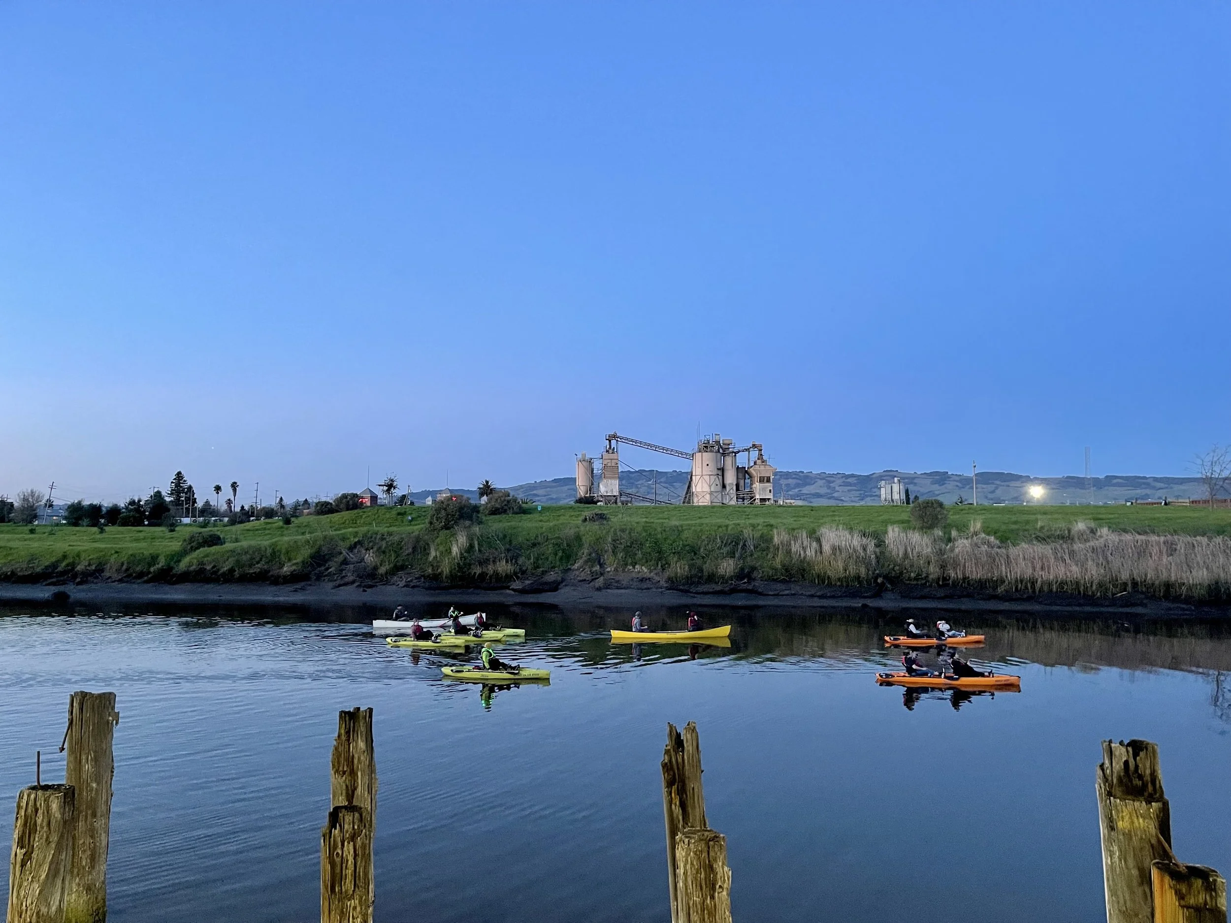 Kayakers paddling on a calm body of water during dusk, with industrial structures and a hill in the background.