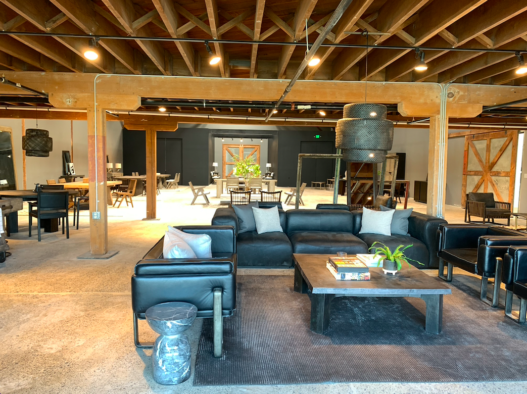 A spacious, industrial-style lounge area with black leather sofas, a wooden coffee table with books and a plant, and a high ceiling with exposed wooden beams and track lighting.