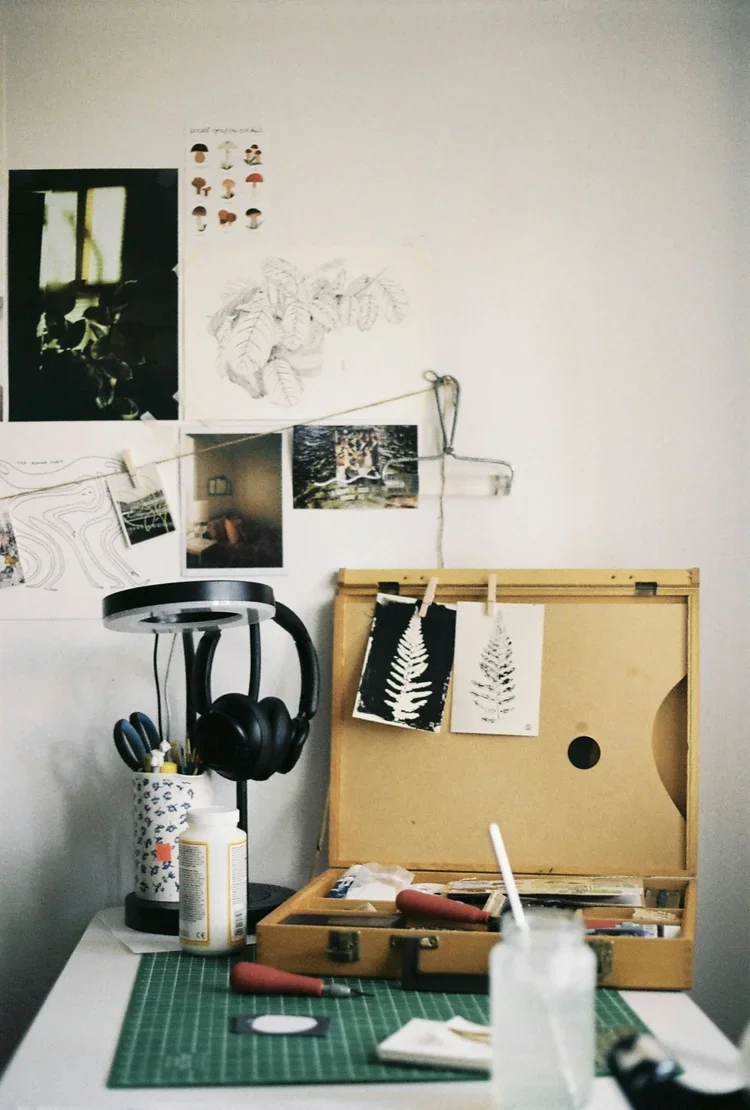 A creative workspace with art supplies and wall decorations. There is a white table with a green cutting mat, a bottle of glue, and art tools. A wooden art case with artwork of leaves, some pinned to a hanging string, and sketches on the wall. A lamp