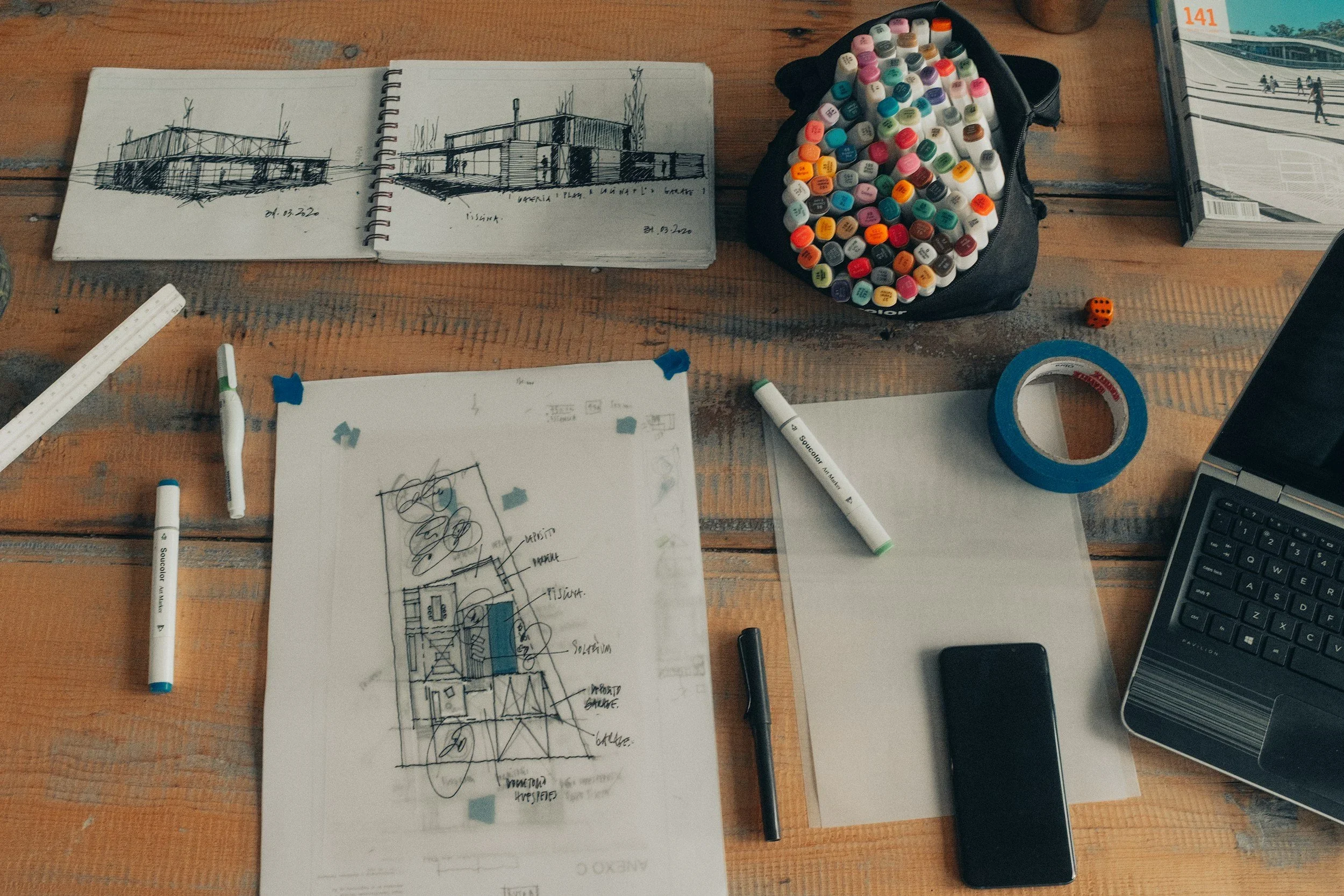 A wooden desk with architectural sketches, colored markers, a roll of masking tape, a black smartphone, a laptop, paper, and an open magazine.