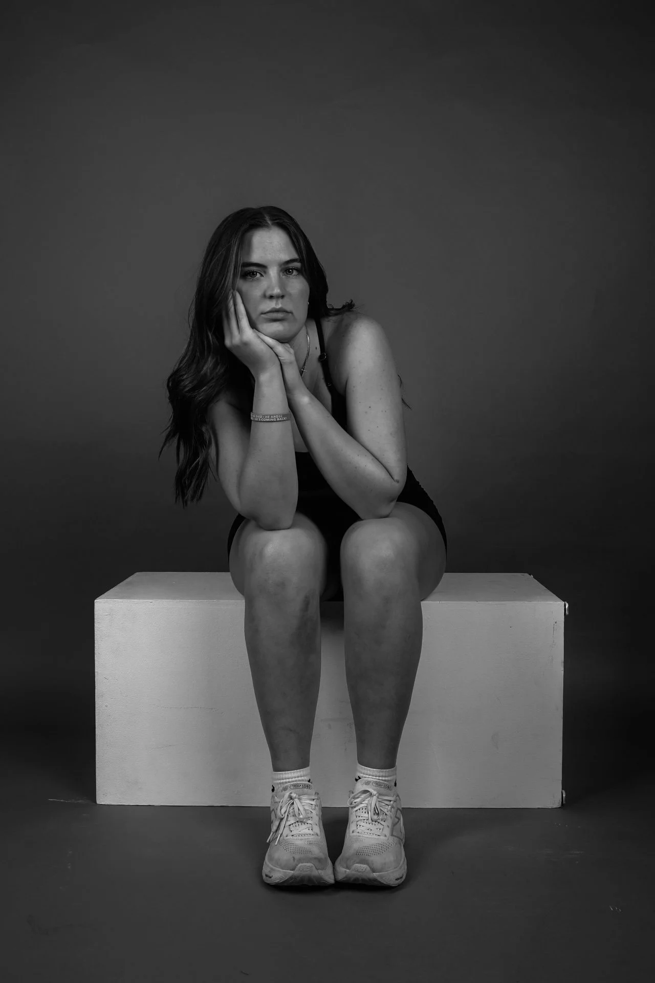 Black and white photo of a young woman sitting on a white box with her chin resting on her hand, looking into the camera with a neutral expression, wearing a tank top, shorts, sneakers, and a wristband.