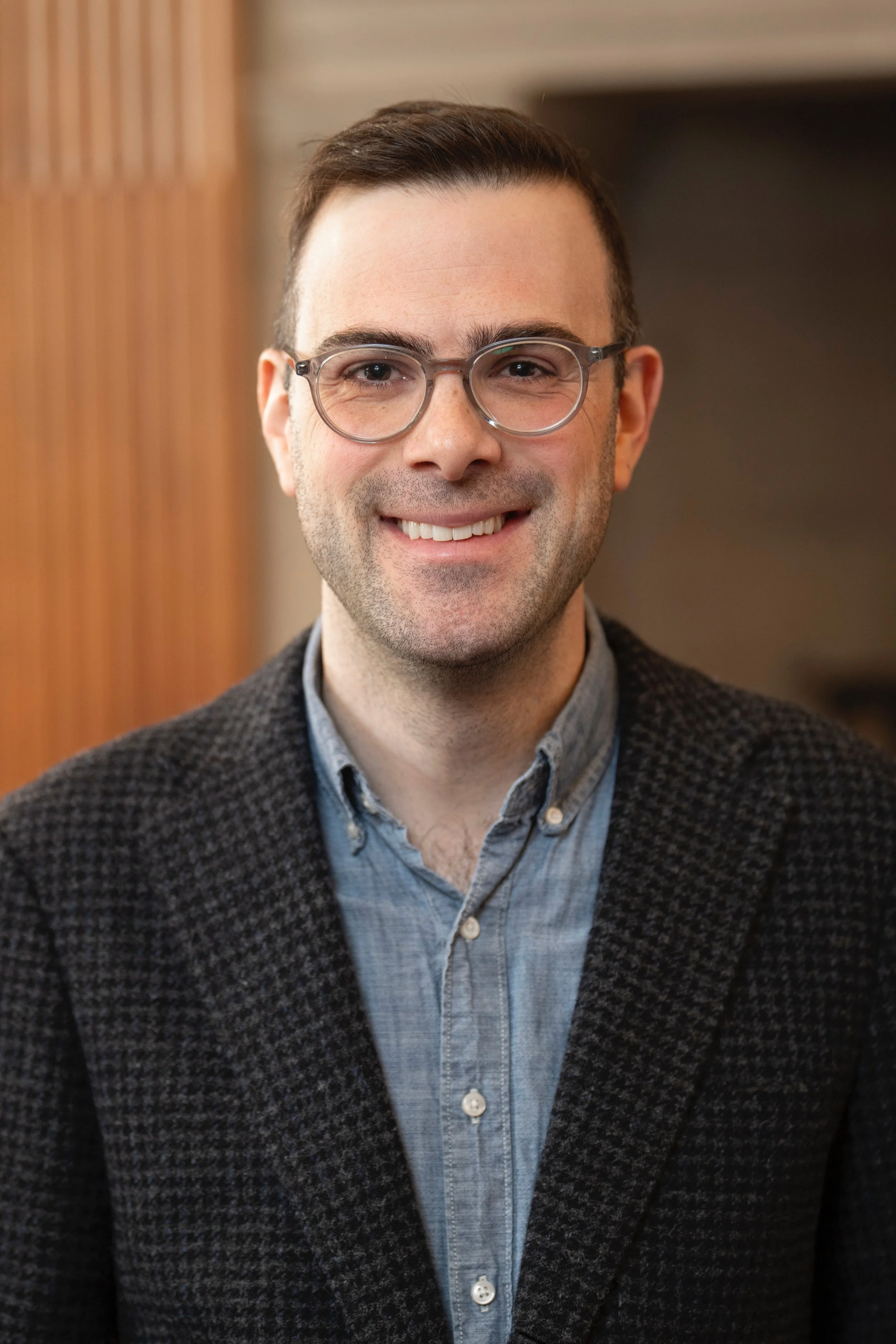 Portrait of a man with glasses, wearing a denim shirt and a dark blazer, smiling, in an indoor setting.