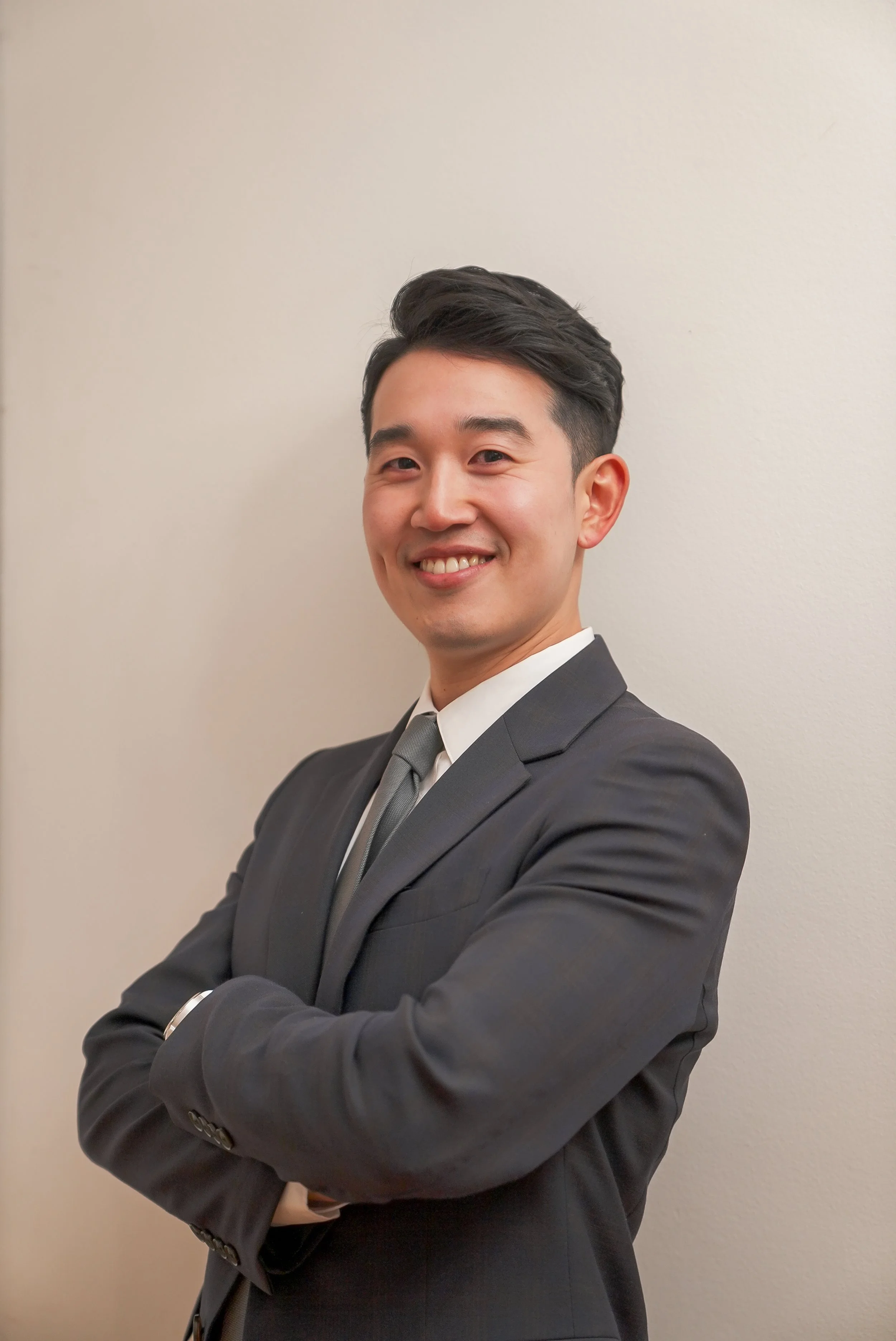 A young man in a dark suit with a gray tie, standing against a beige wall, smiling with arms crossed.