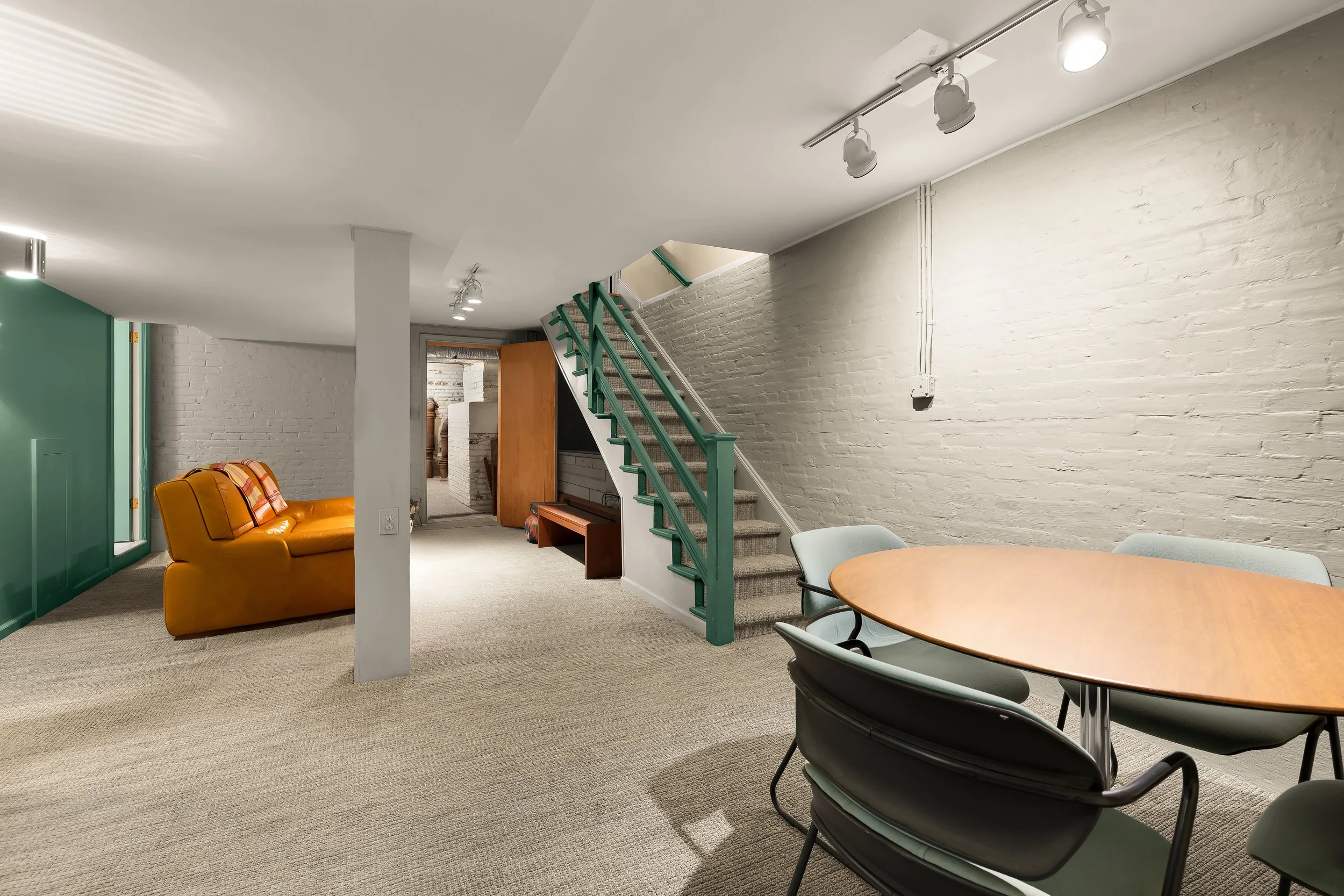 Interior of a basement room with a round wooden table, light green chairs, a yellow sofa, a staircase with a green railing, and various furniture pieces against white brick walls and neutral carpeted flooring.