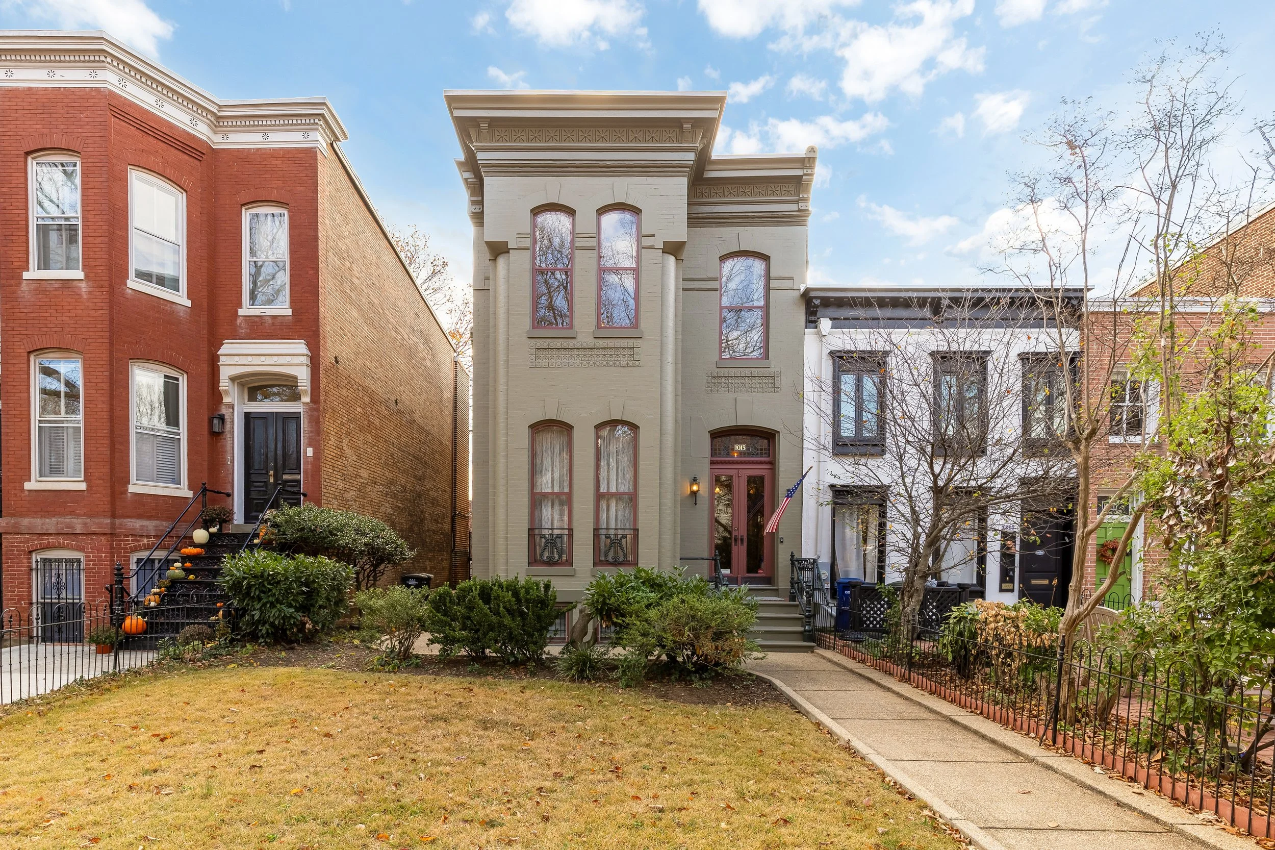 Front view of a three-story townhouse with beige facade, large arched windows, and stairs leading to the front door, next to two similar houses, with a small front yard and leafless trees.