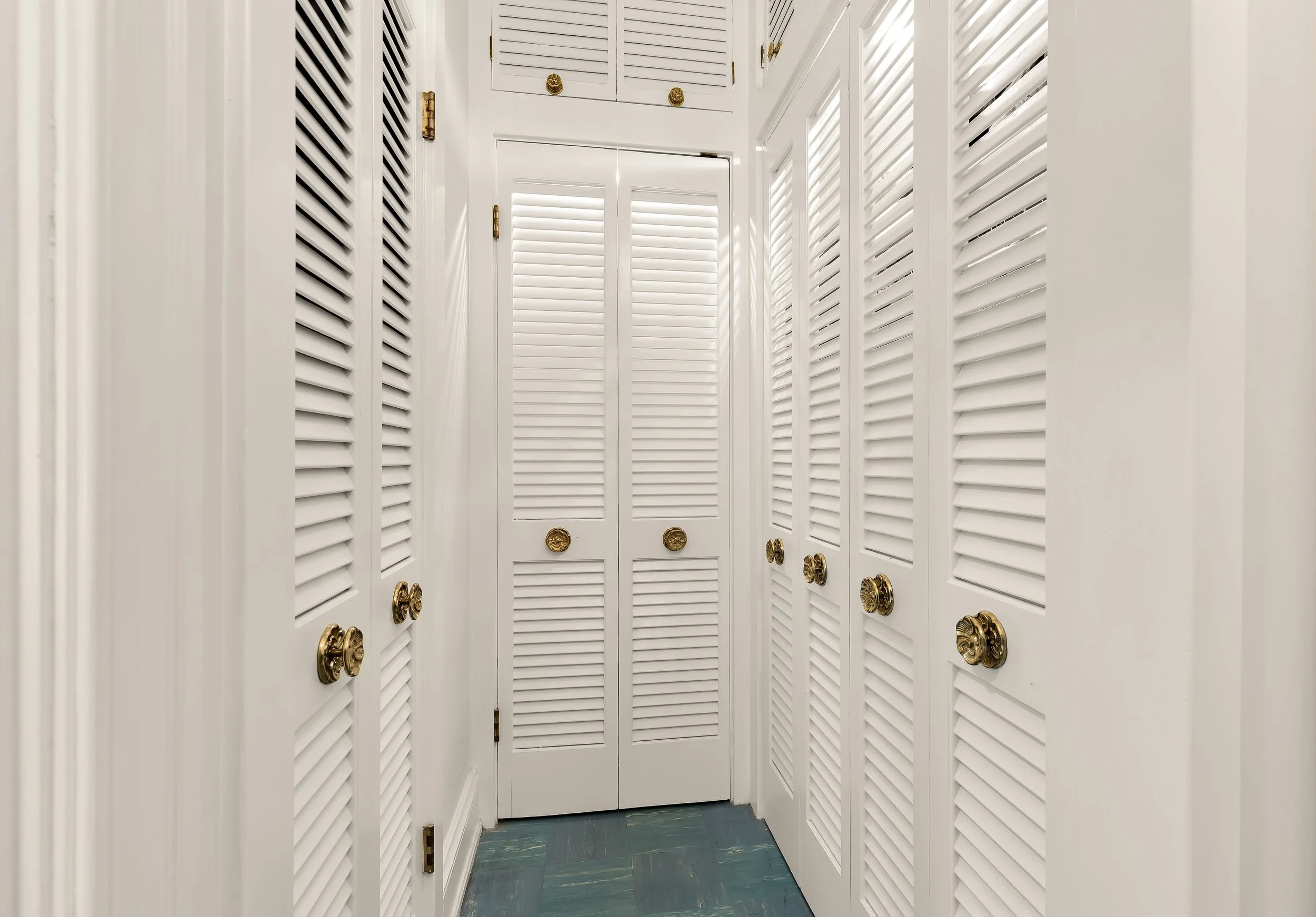 A hallway with white louvered closet doors and gold knobs, with light coming through the slats, and a tiled floor.