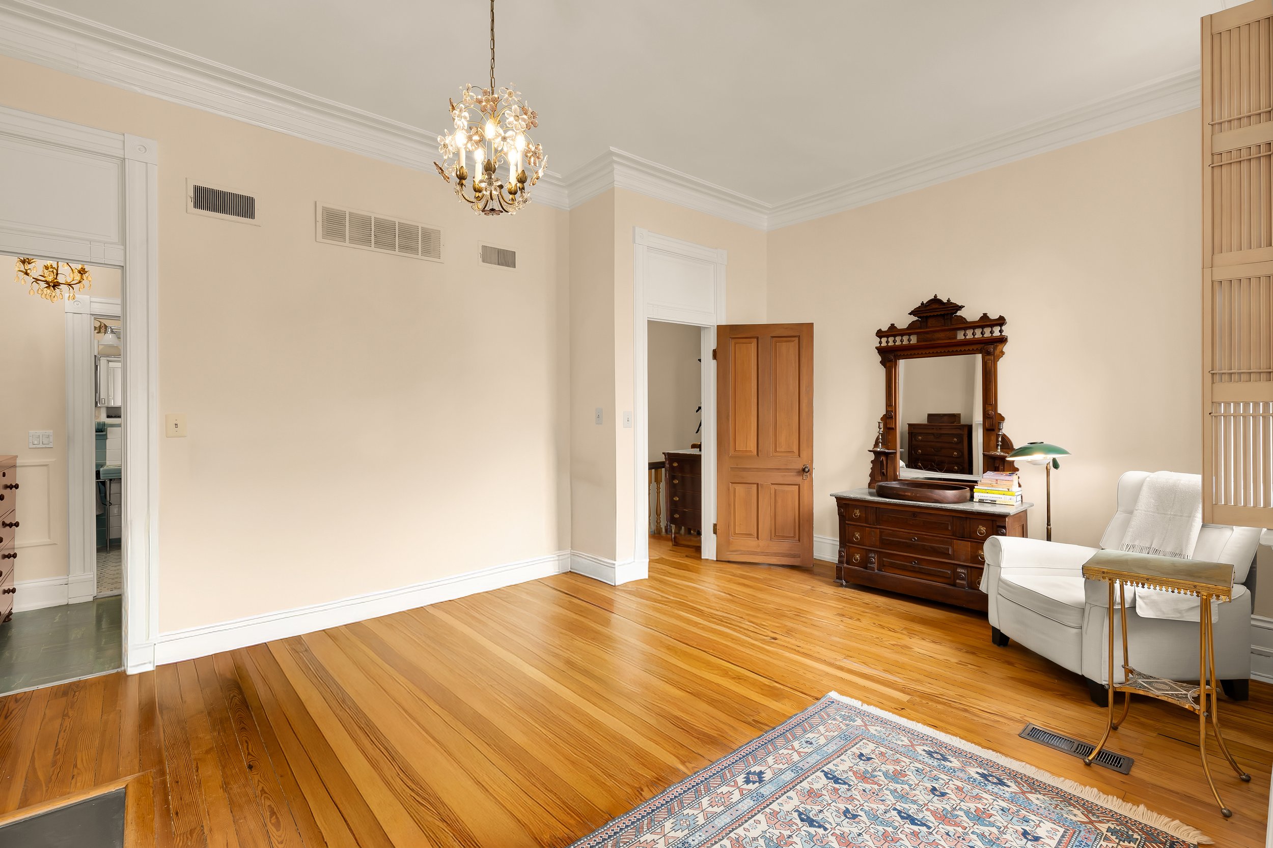 Living room with hardwood floors, beige walls, vintage wooden furniture including a dresser with a mirror, white armchair, floor lamp, and a chandelier hanging from the ceiling.