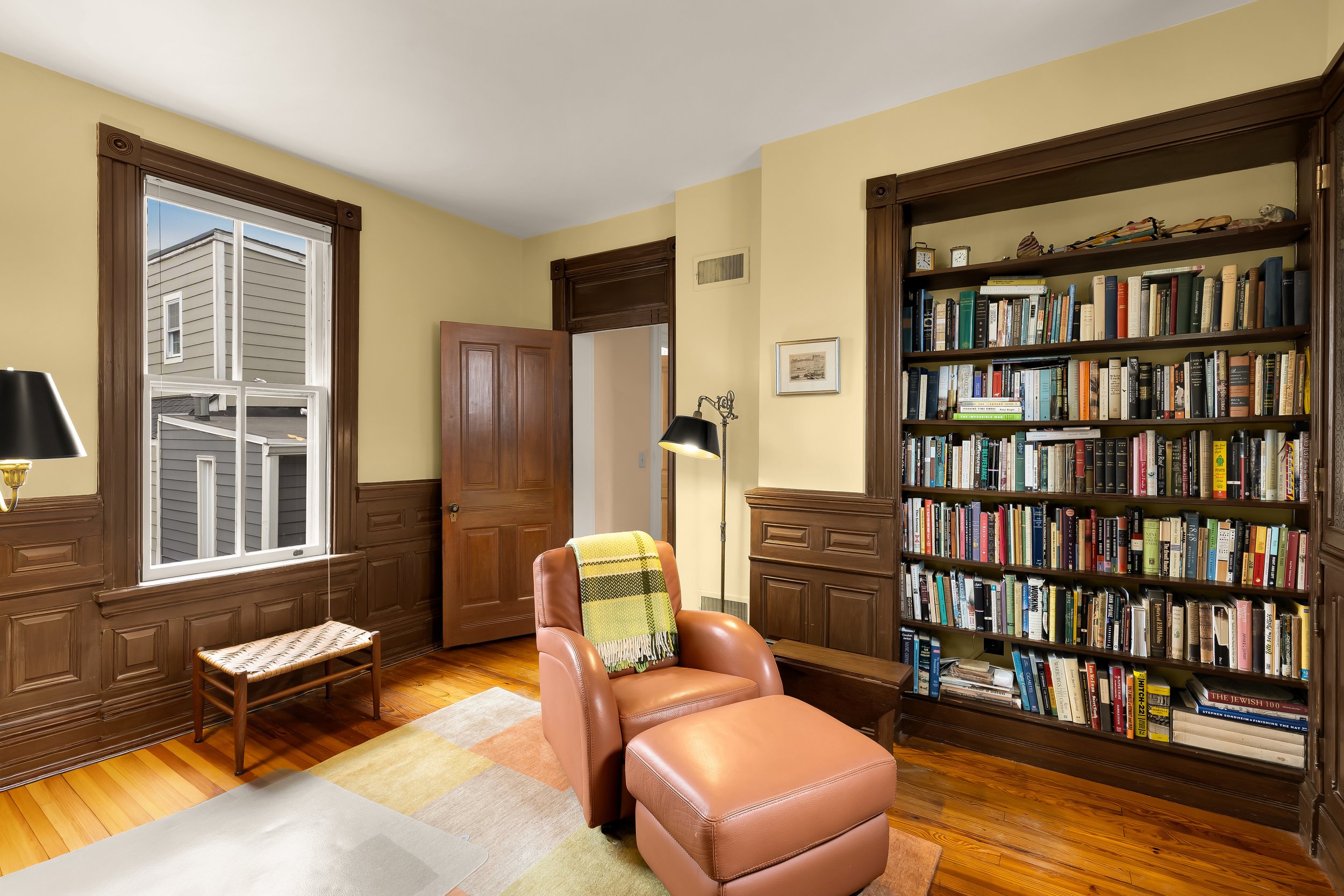 A cozy reading nook with hardwood floors, a pink leather armchair and matching ottoman, a floor lamp, a large bookshelf filled with books, a window with wood trim, and a small bench under the window.