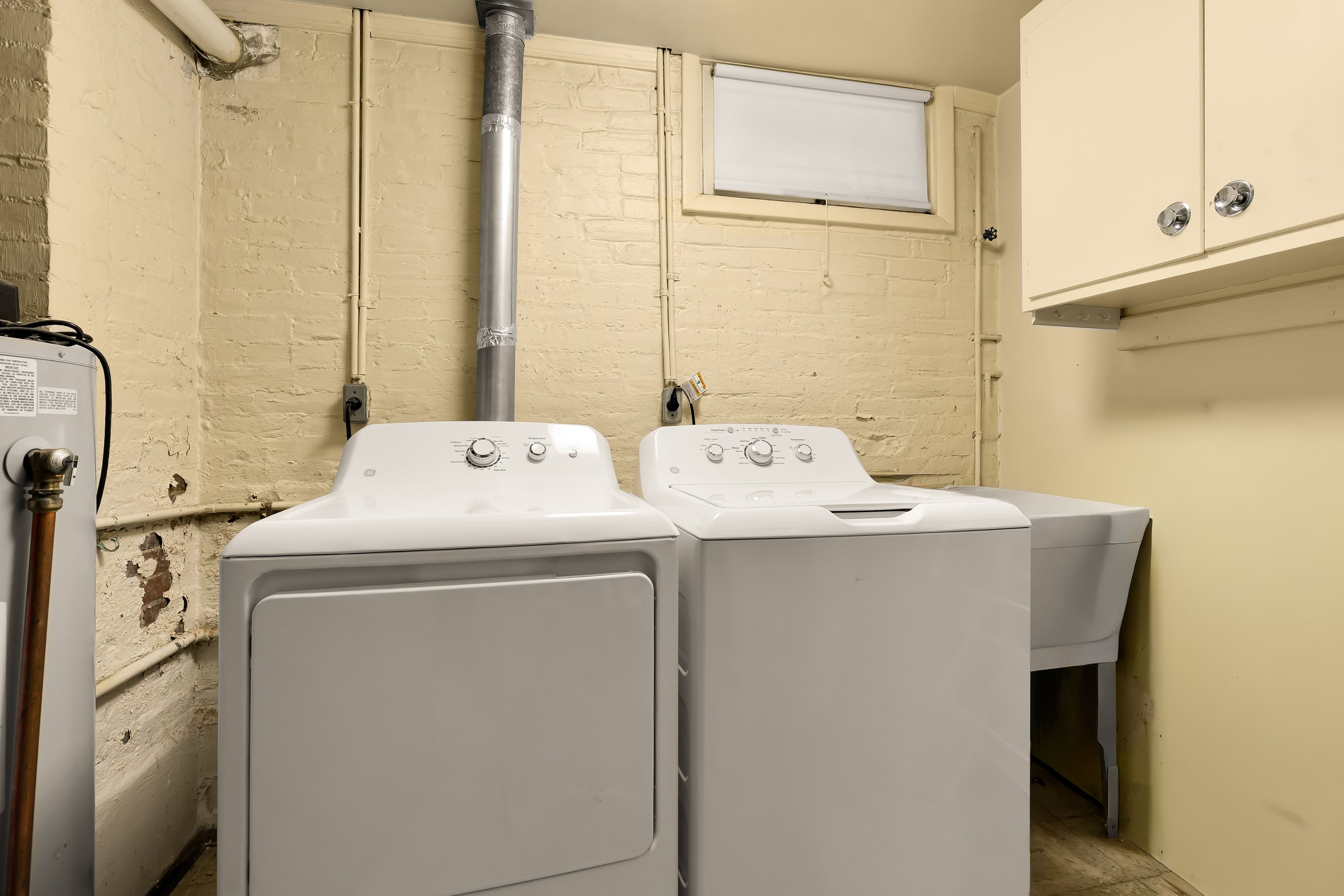 Laundry room with a washing machine and a dryer in front of a brick wall with pipes and a small window.
