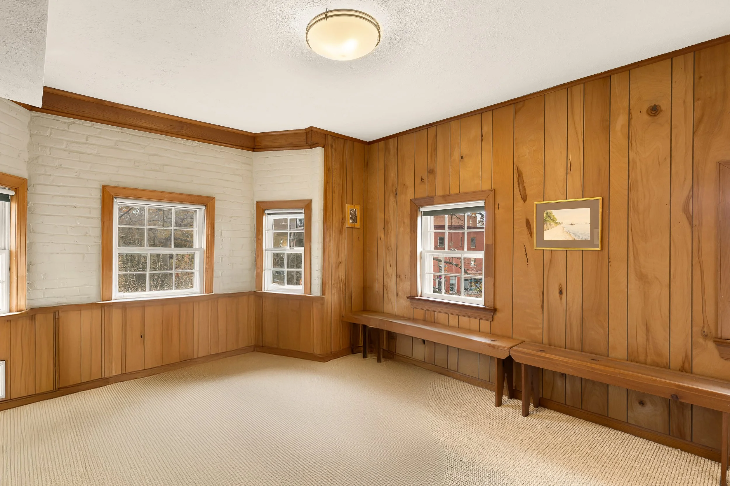 A room with wood paneled walls, three windows with white trim, beige carpet, and a ceiling light fixture.