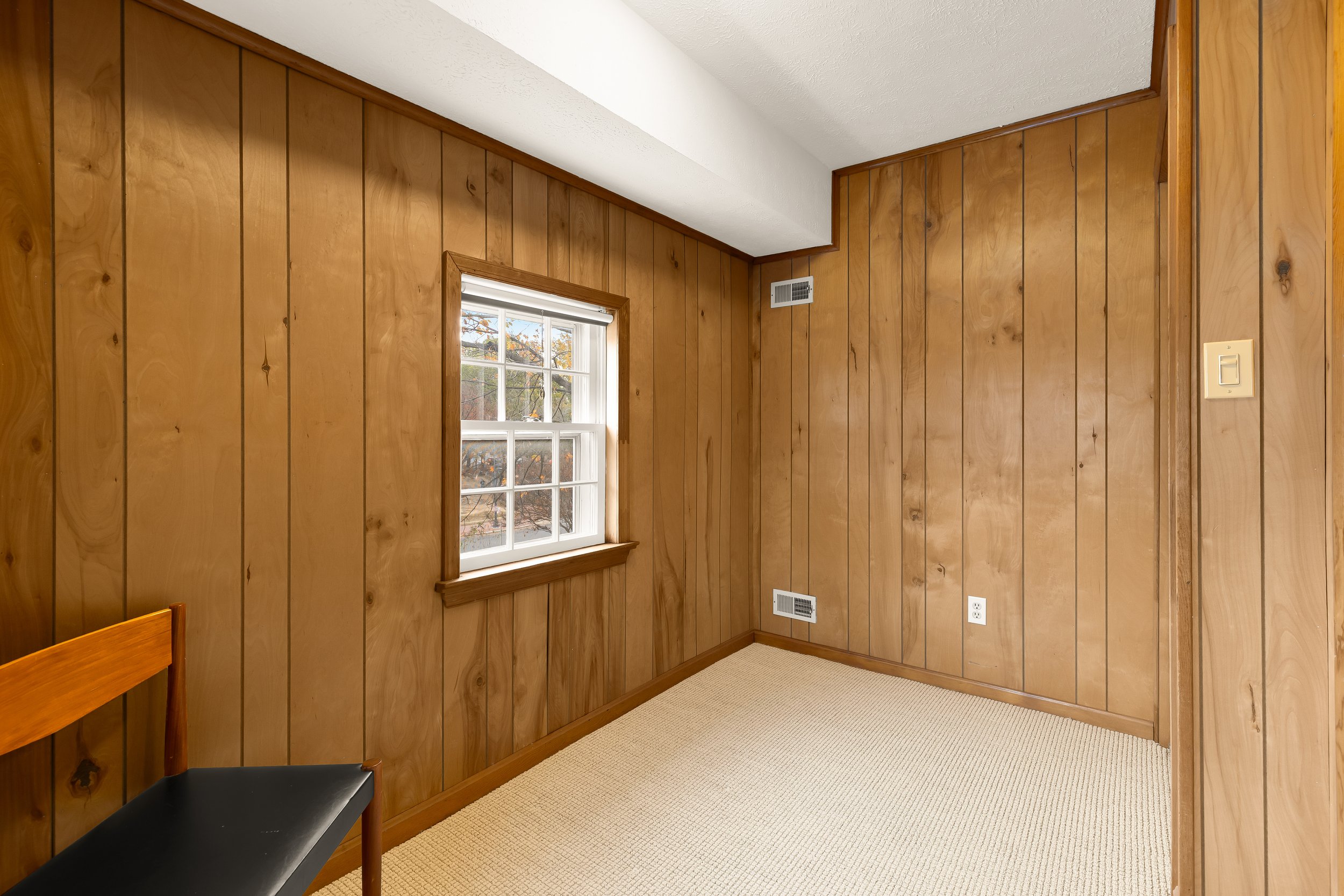 Empty room with wood-paneled walls, beige carpet, a window with white frame, and a chair with a black seat cushion.