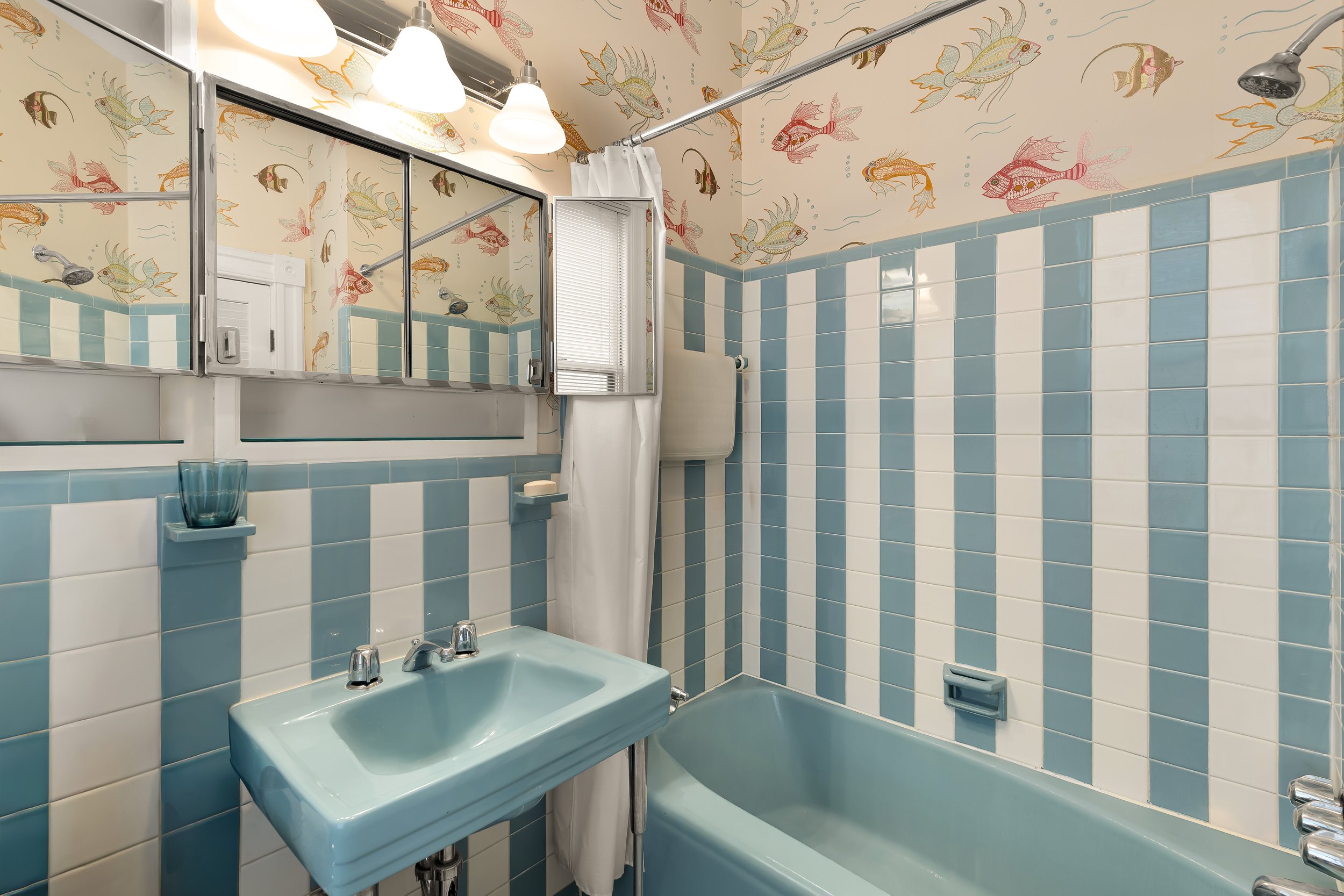 A vintage bathroom with blue and white tiled walls, fish-patterned wallpaper, a blue sink, a blue bathtub, a small mirror, a soap dish, and a window with blinds. There are white light fixtures above the mirror.