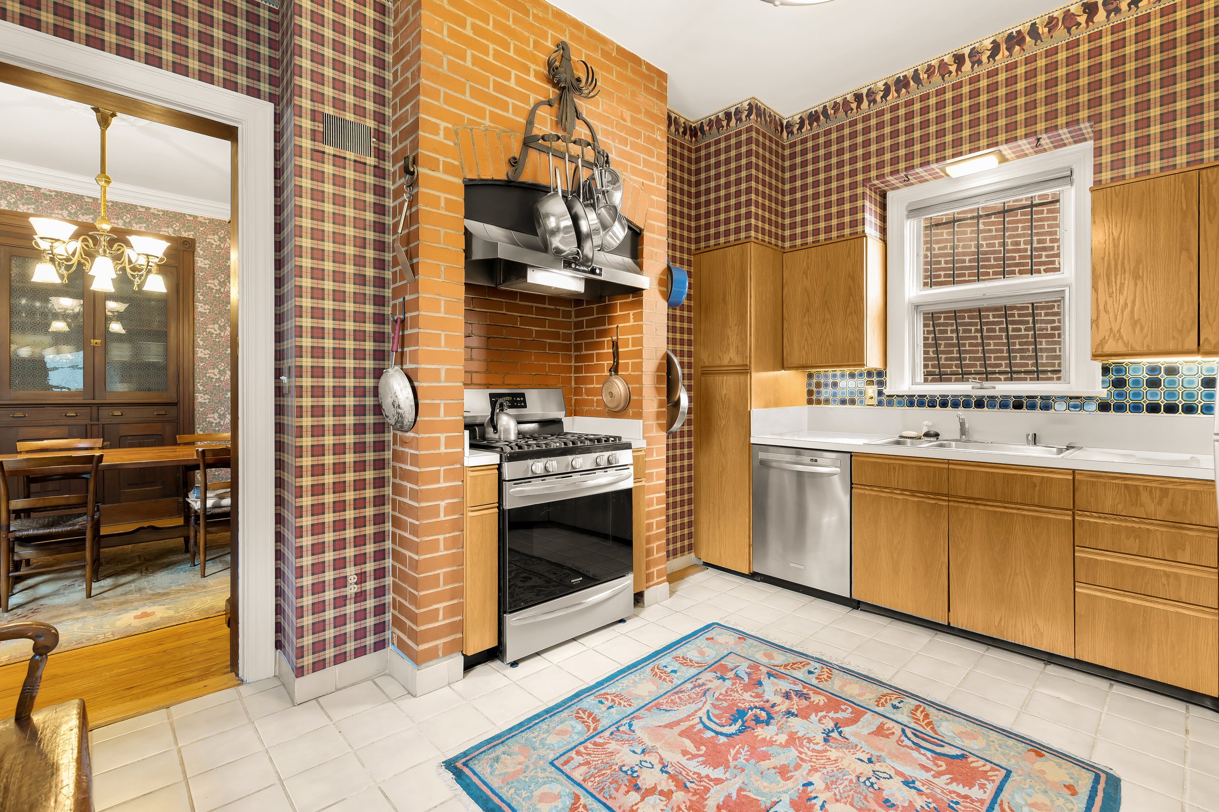 Kitchen with brick wall, wooden cabinets, stainless steel appliances, window, patterned wallpaper border, and a colorful rug on tile floor.