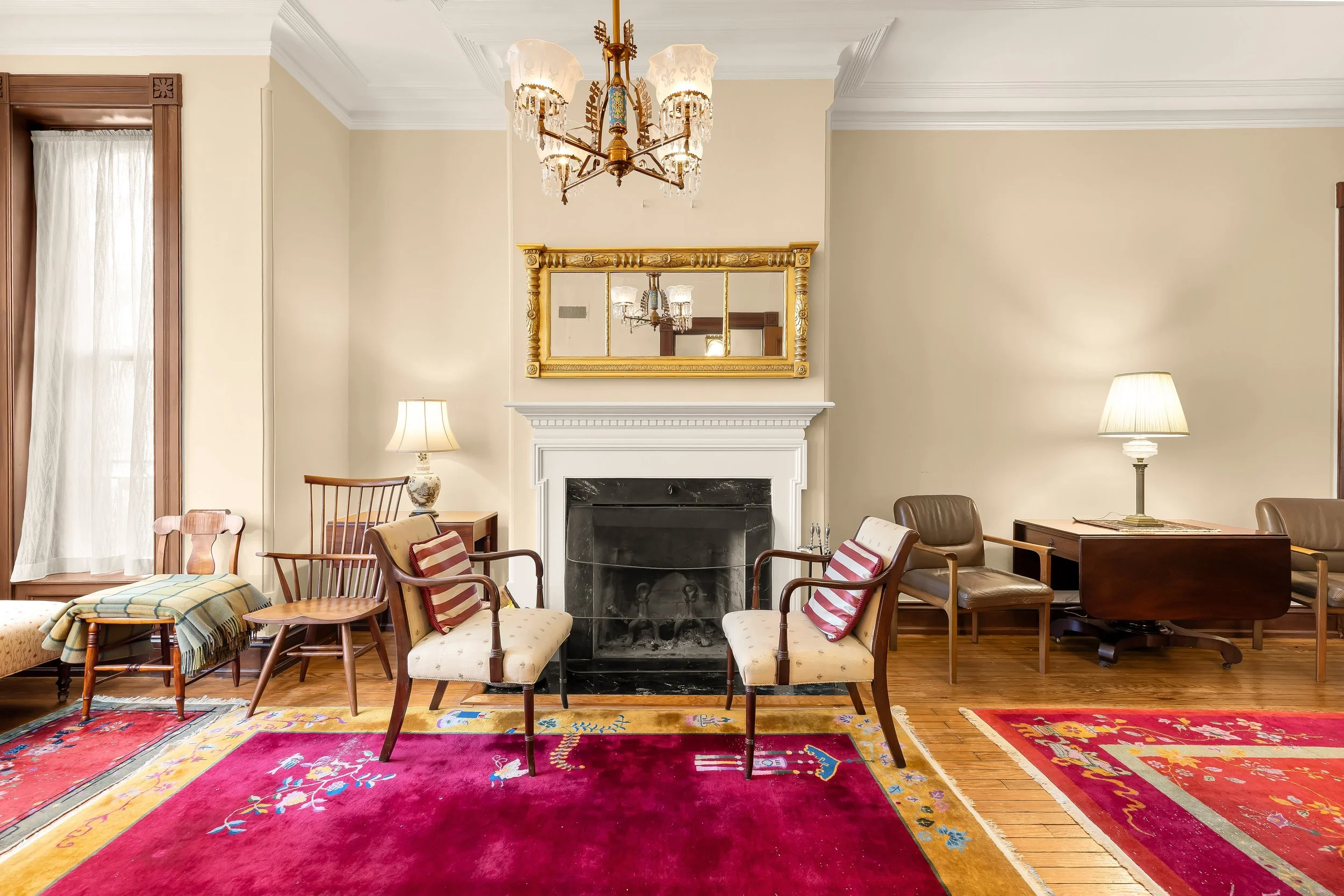Living room with a fireplace, multiple vintage chairs, side tables with lamps, and colorful rugs on hardwood floor, decorated in a classic style.