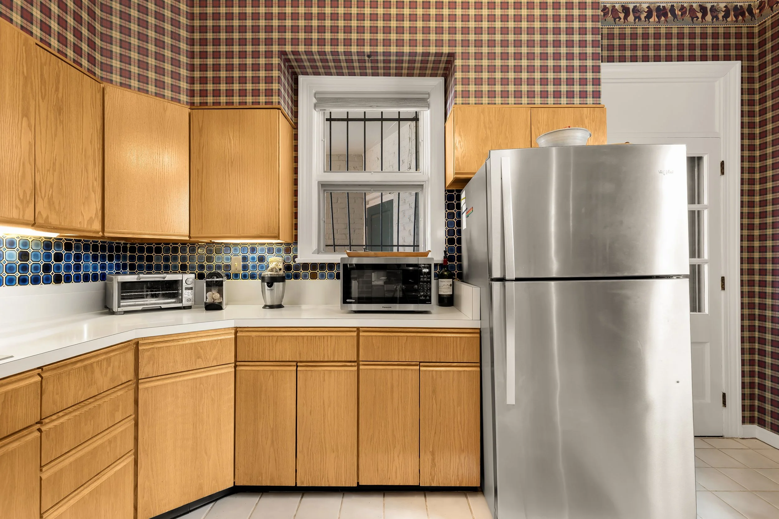 Kitchen with wood cabinets, a colorful tiled backsplash, a stainless steel refrigerator, and a window with bars.
